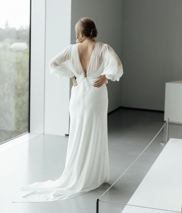  A midsized bride standing with her back to the camera in front of a large window. She is wearing a long-sleeved, sheer gown with a V back. 