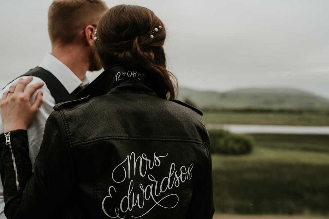  A bride and groom are pictured looking out to the countryside in the distance with their backs to the camera. The bride is wearing her black leather Jacket with her personalised Mrs painted in white calligraphy across the back. She is wearing the co