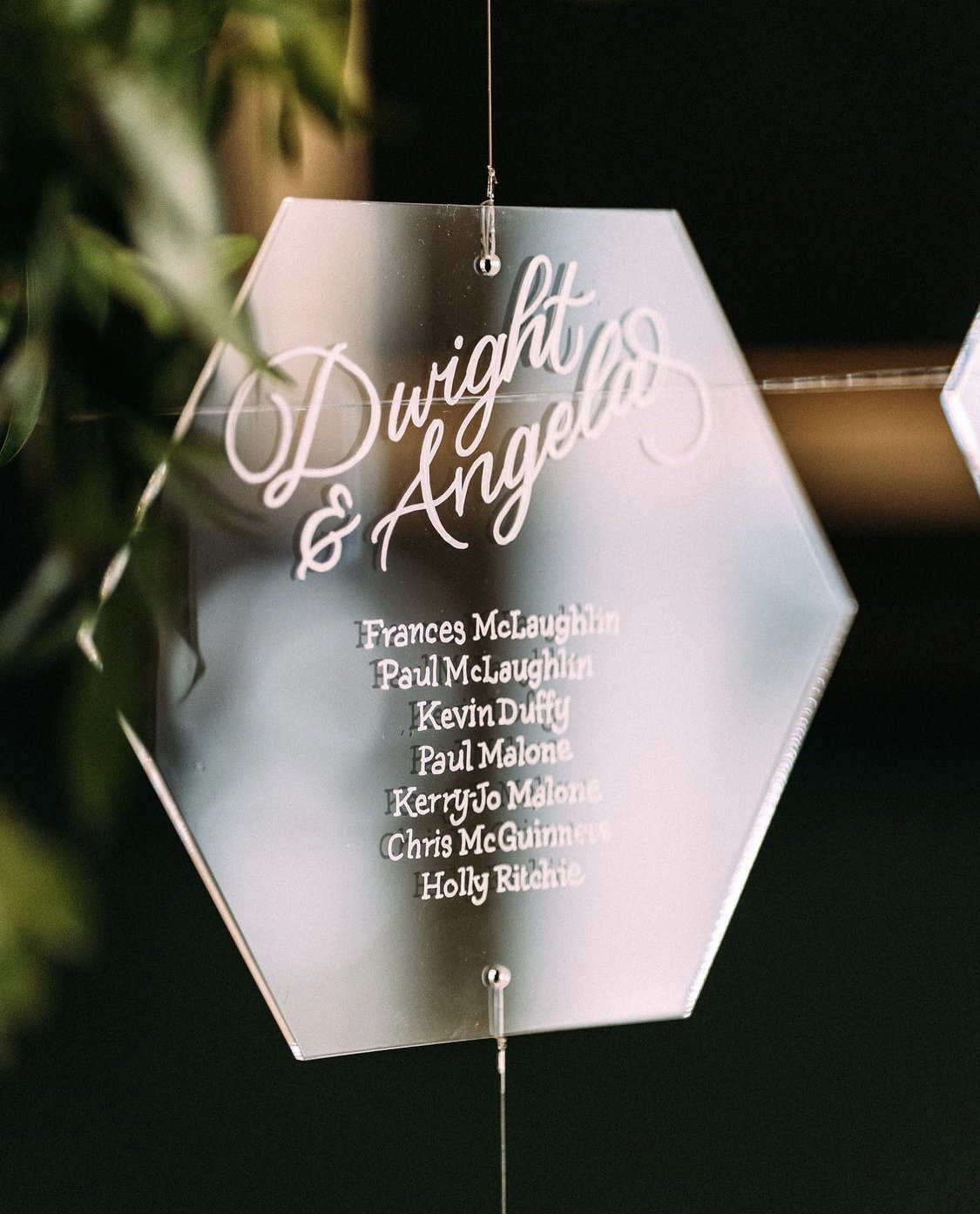  A hexagonal hanging acrylic table plan is hung on fishing wire. The bride and groom’s names are at the top with wedding guests names for that table underneath. The acrylic plan is displayed within green leaves which can just be seen to the left of t