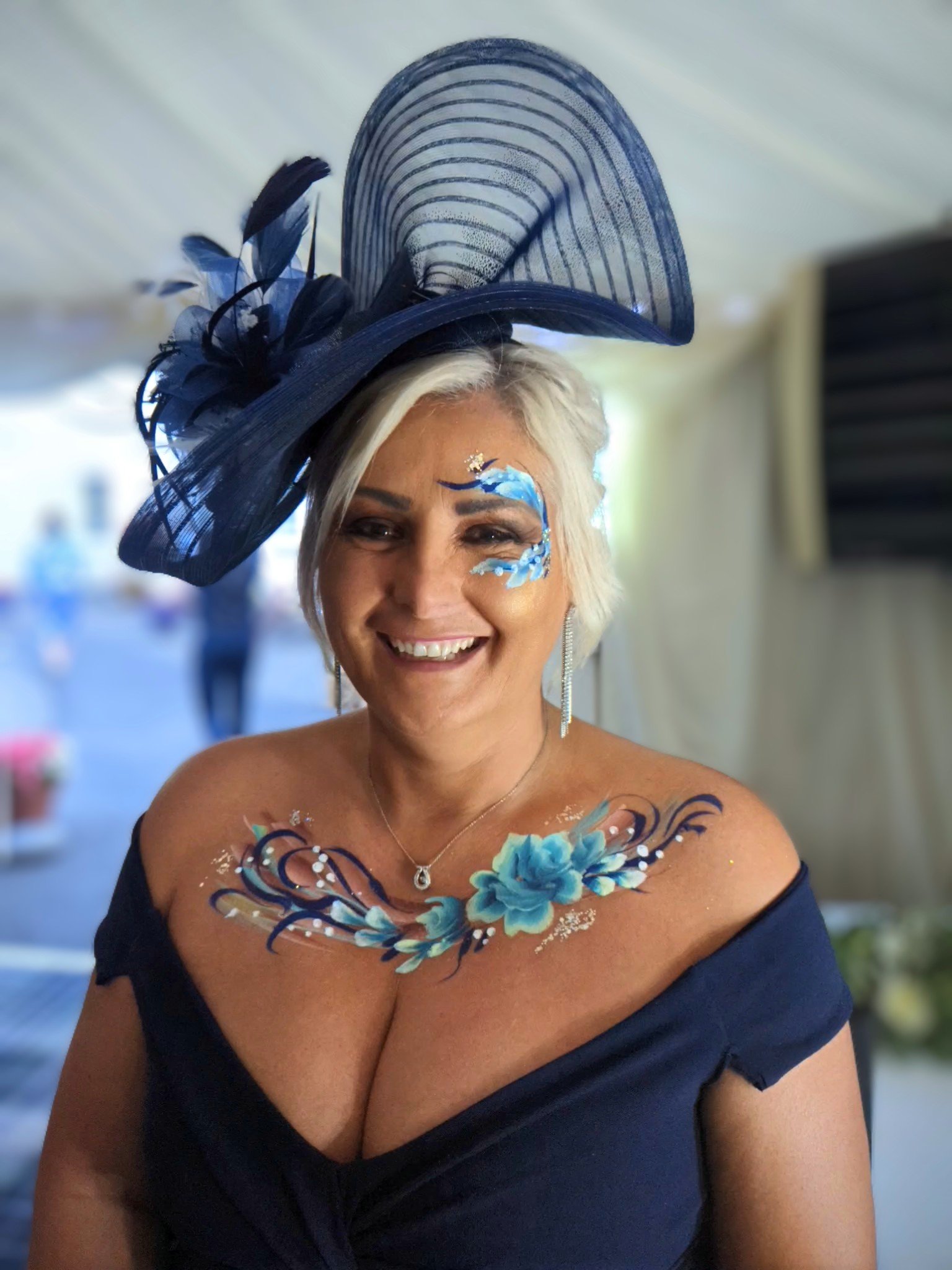  Wedding guest is wearing a navy off the shoulder dress with navy fascinator. Gorgeous blue swirl facepaint is around the eye area with glitter and further hand painted florals that compliment the wedding guests outfit across her chest.  