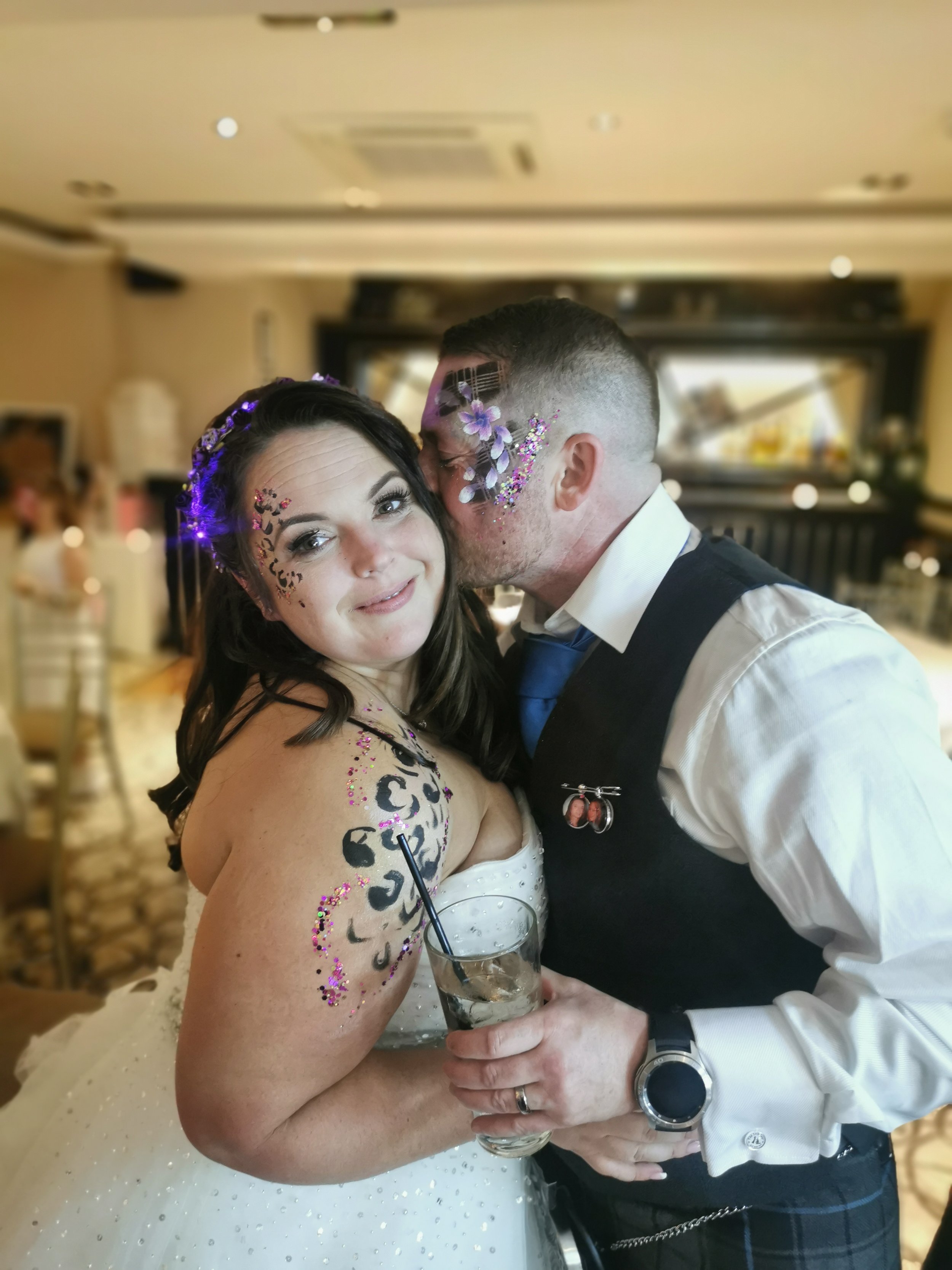  Bride wearing a white strapless wedding dress is facing the camera with glitter and facepaint around her eye and further glitter and hand painted artwork from her shoulder and down her arm. The groom is stood kissing the bride on the cheek and displ