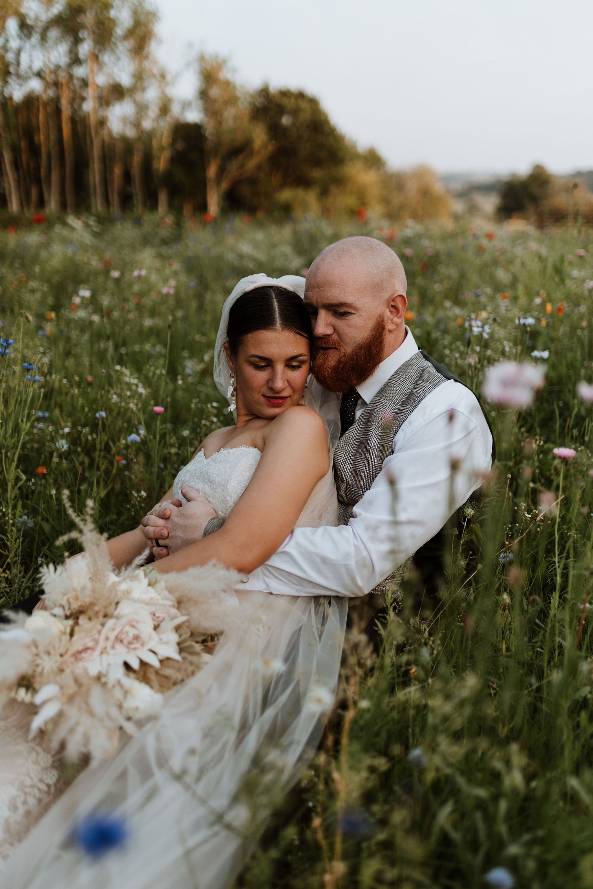  The bride and groom are sat together in a meadow. The bride is sat with her back against the groom who has his arms wrapped round her whilst their heads are touching. The bride is wearing a white strapless wedding dress, with a white veil behind her