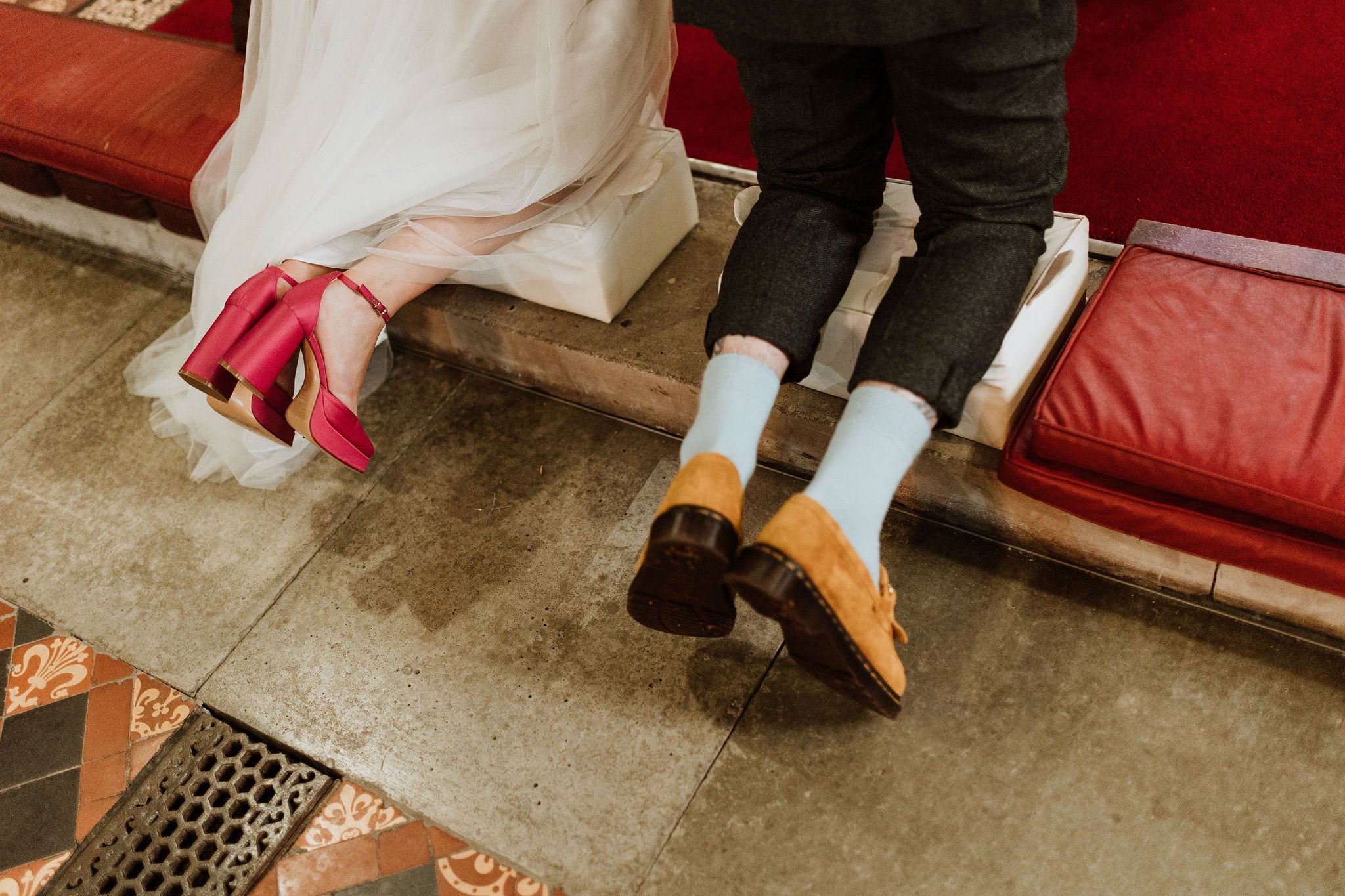  The bride and groom are kneeling down on white cushions placed on the stone church floor. You can see the brides bright pink platform heeled shoes with a pink ankle strap and you can just see the material from her long white wedding dress. To the ri