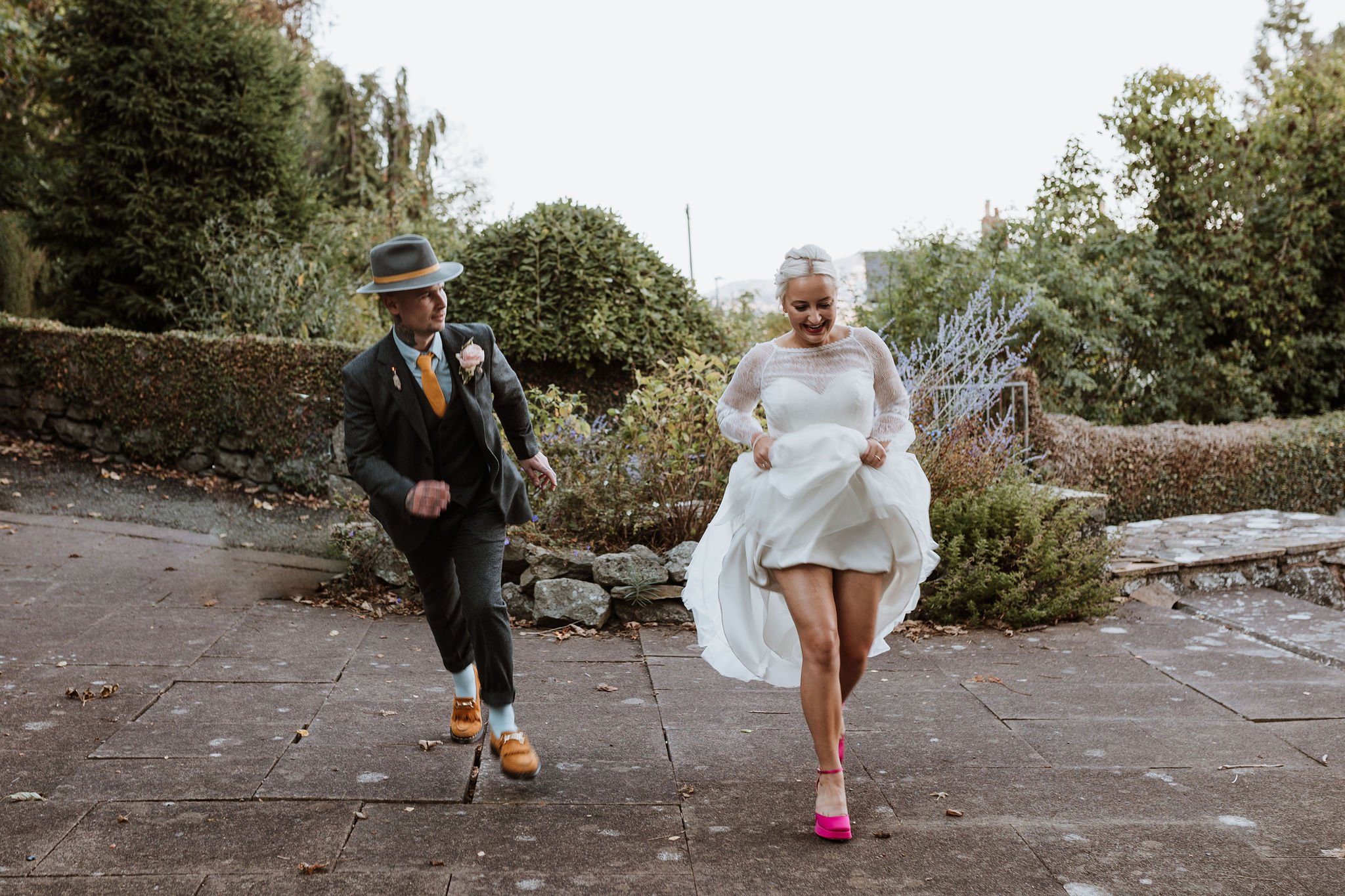  The bride and groom are captured next to each other outside mid dance.   The bride has her long white wedding dress hitched up above her knee allowing herself to dance and is looking down towards her bright pink platform shoes.   The groom is to her