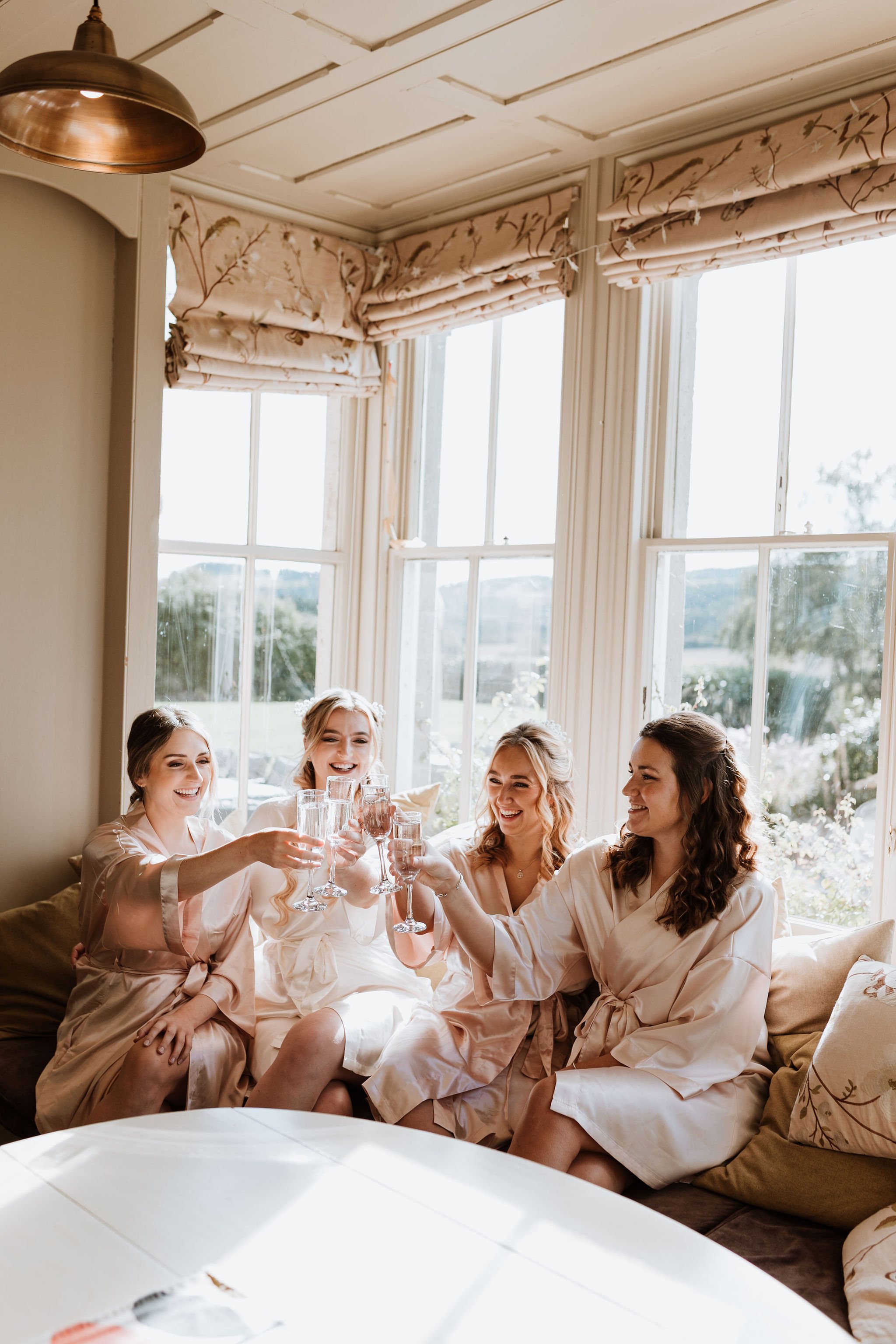  The bridal party are sat wearing cream silk dressing gowns, chinking champagne flutes in a bright room with a big bay window behind them.  