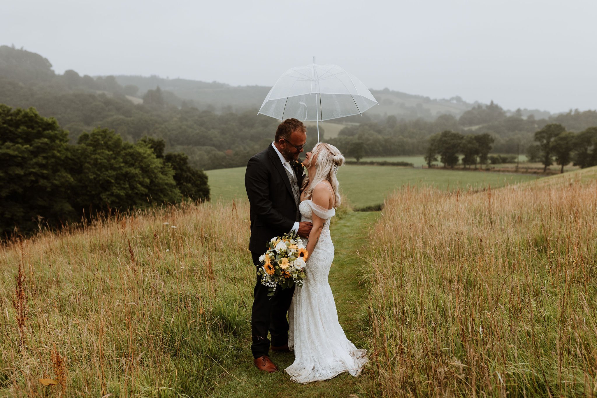  Stood on a mowed path of grass, surrounded by fields and trees the bride and groom are stood gazing into each others eyes under a clear umbrella which they are holding up over themselves.   The groom is wearing a dark suit with a checked waistcoat a