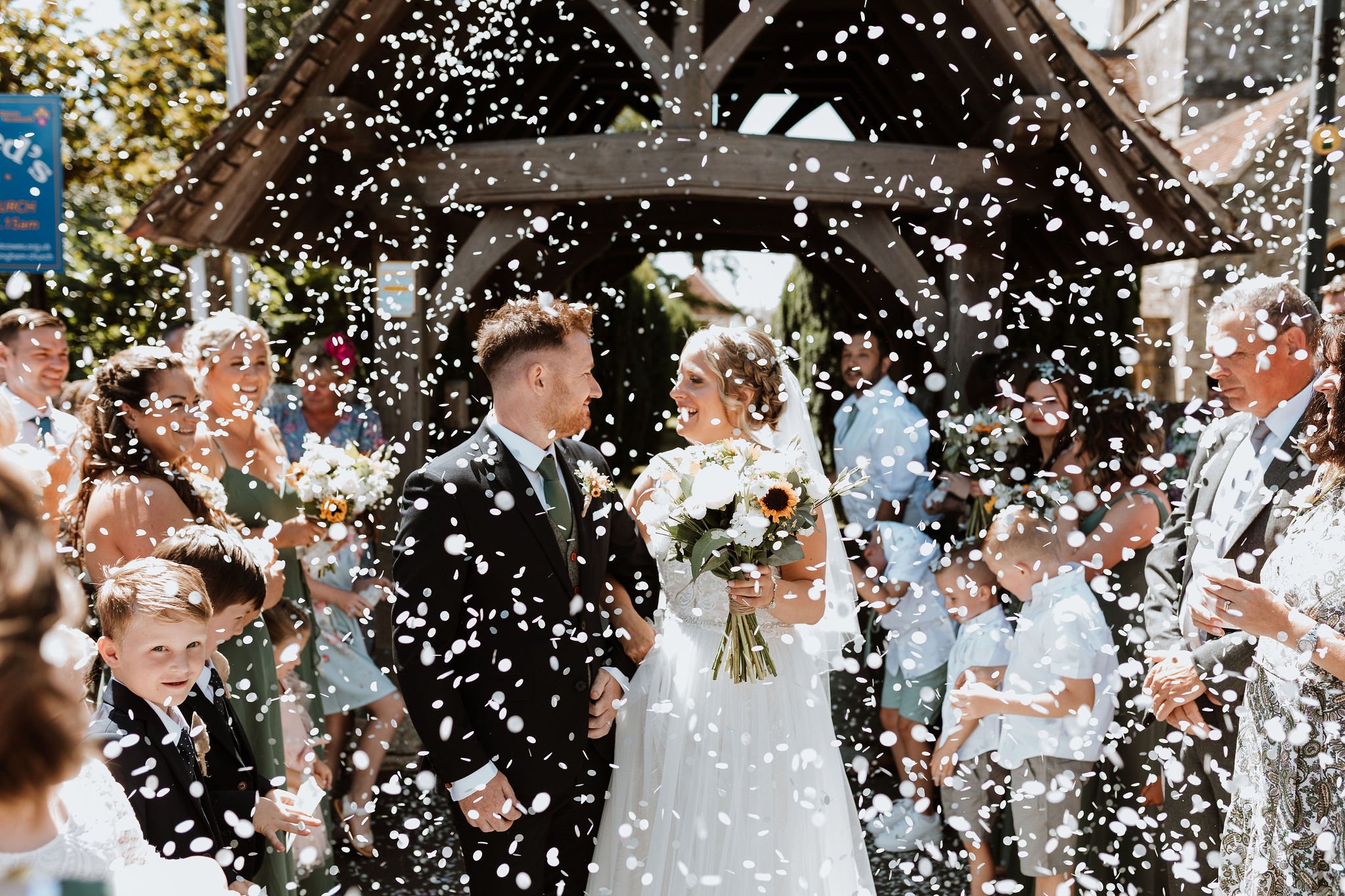  Bride and groom are looking at each other after leaving the church with their wedding guests on either side throwing white confetti over them. The groom is wearing a dark suit with a tweed waistcoat, white shirt and green tie.  The bride is wearing 