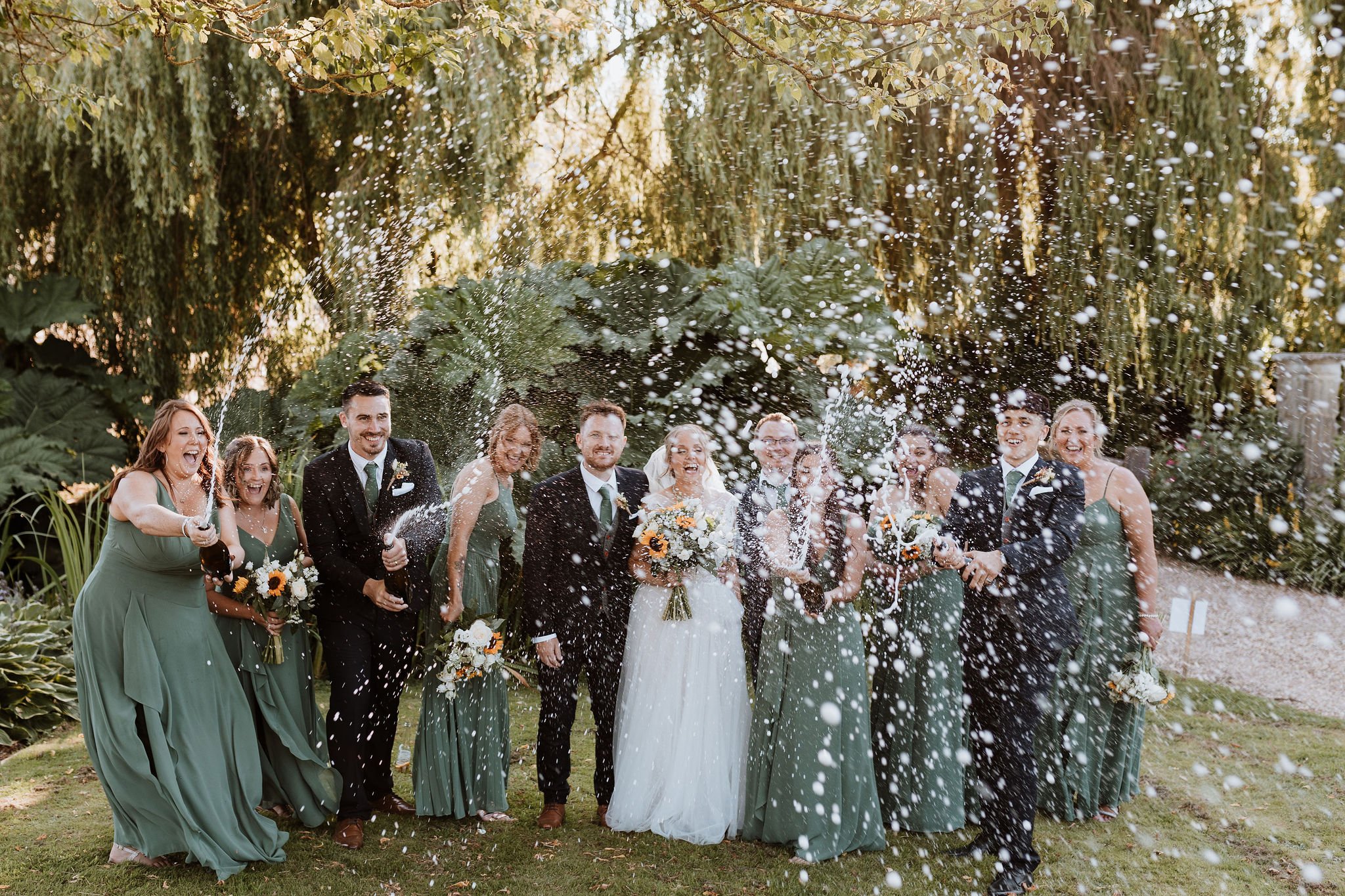  The wedding party are stood on a lawn with large foliage behind them. Various bridesmaids and groomsmen are spraying champagne towards the photographer with the bride and groom stood in the centre laughing and smiling. The bride is wearing a long wh