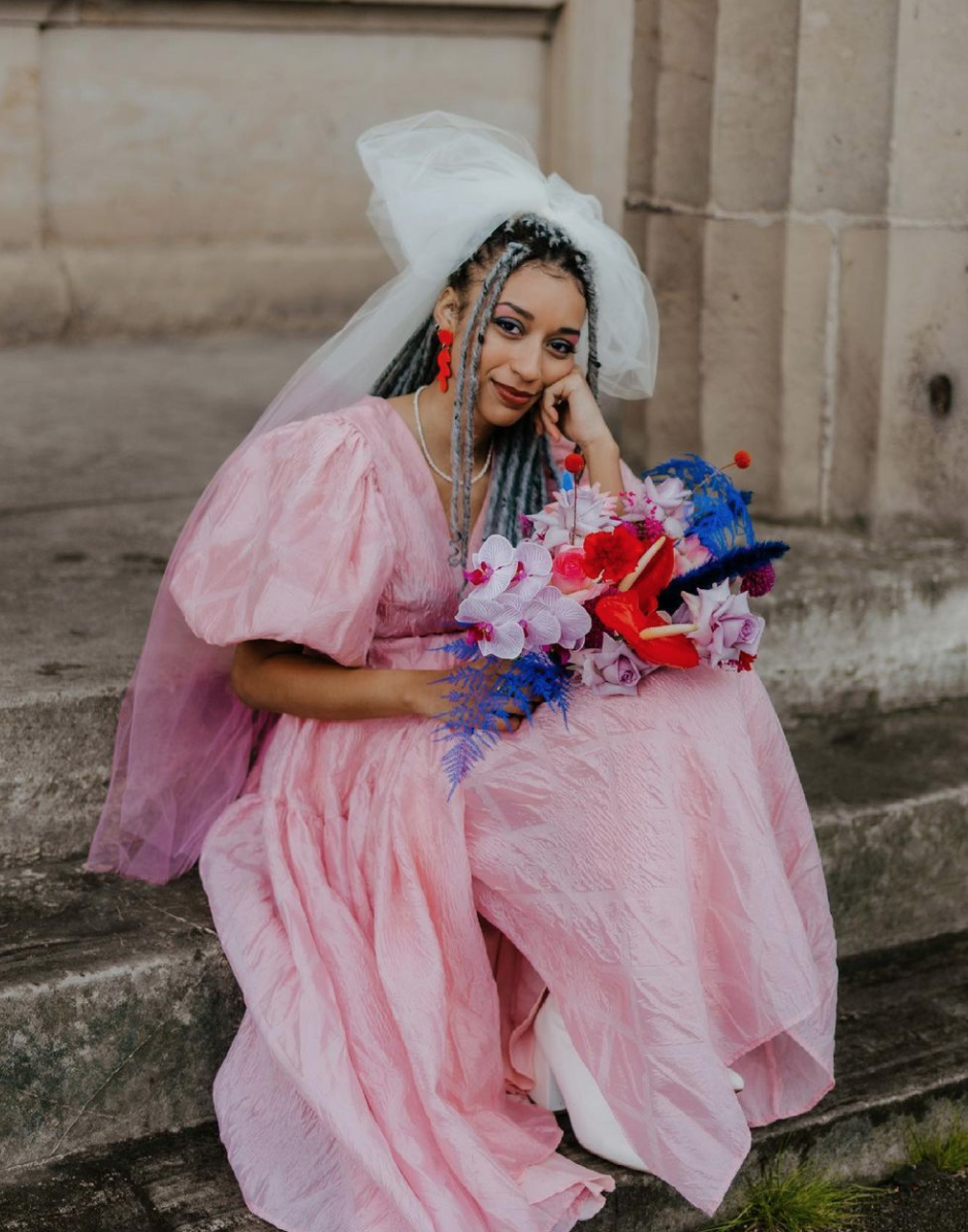  Bride wearing a beautiful long, pink wedding dress with puffball sleeves is sat on concrete steps with her head resting on her hand. She is wearing an awesome white veil in her hair which has been dipped pink at the end. The wedding florals which ma