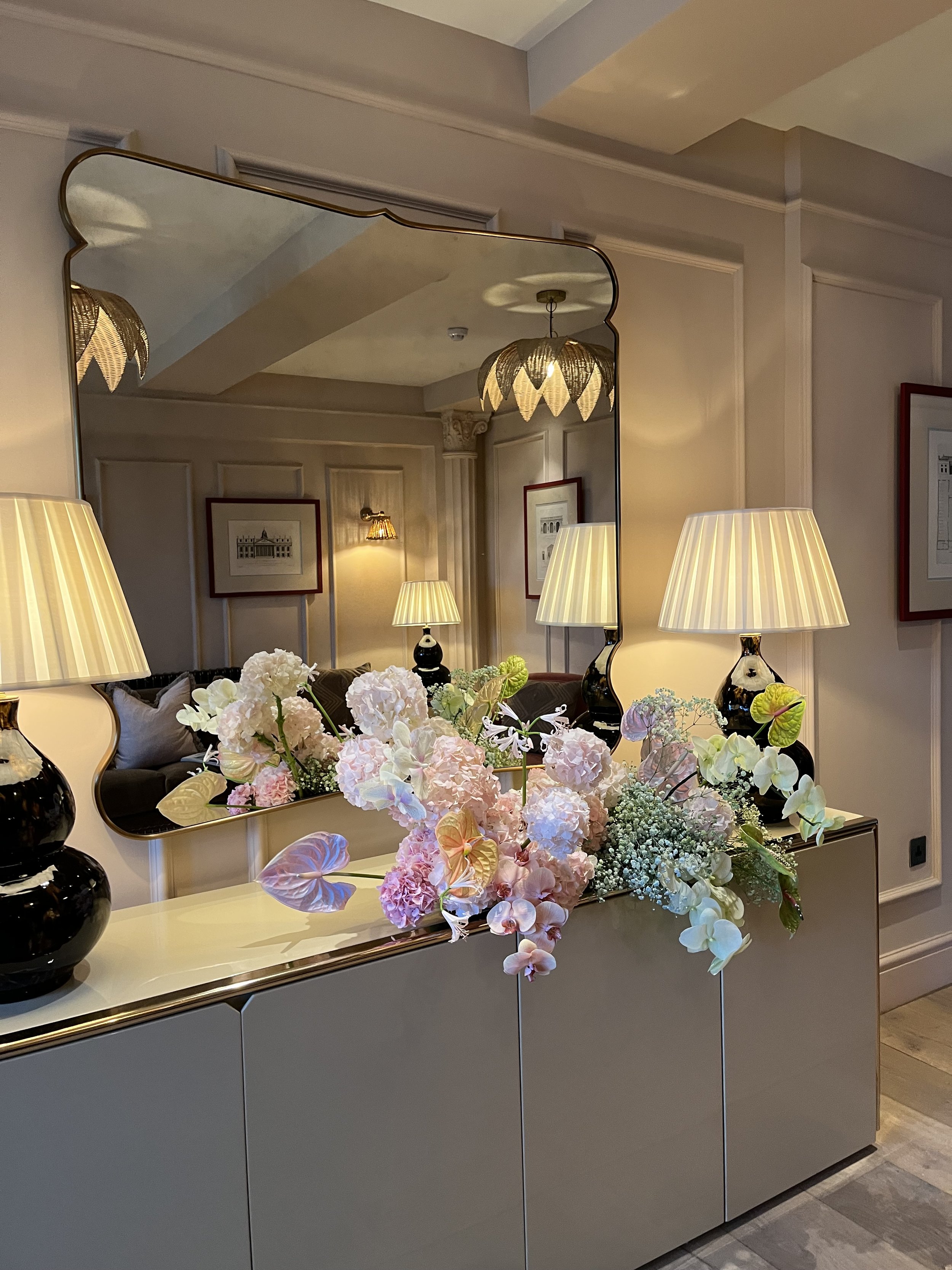  Beautiful wedding flowers have been placed on the side in a hotel room in front of a mirror. The stunning wedding floral installation is created using large blooms in soft pinks and whites alongside gypsophila and leaves, all placed at different hei
