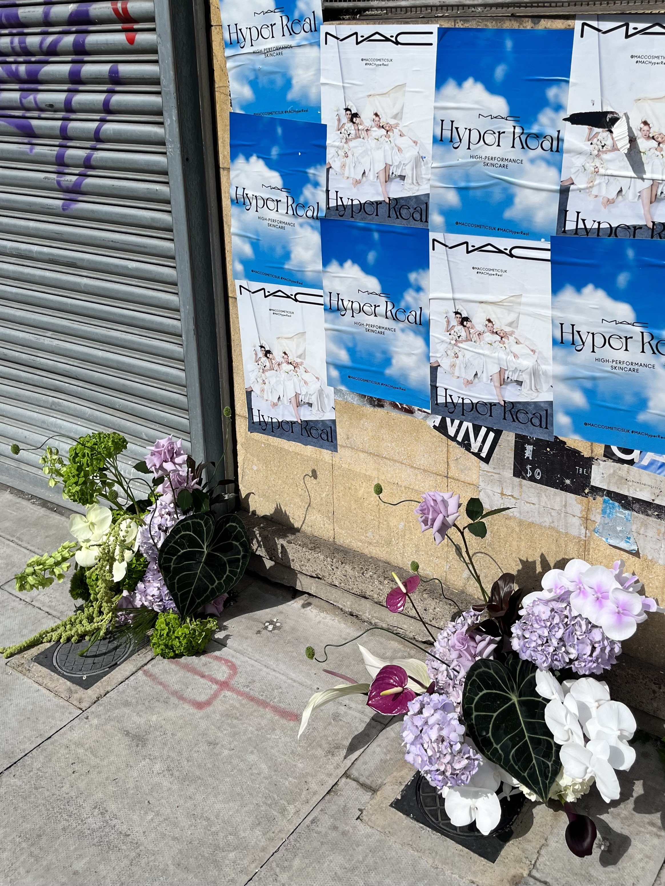  MAC make up posters stuck on a wall outside next to a metal shutter is the backdrop for two wedding floral displays which have been placed on the pavement. The bold wedding blooms in purple and white and the large green leaves create a striking disp