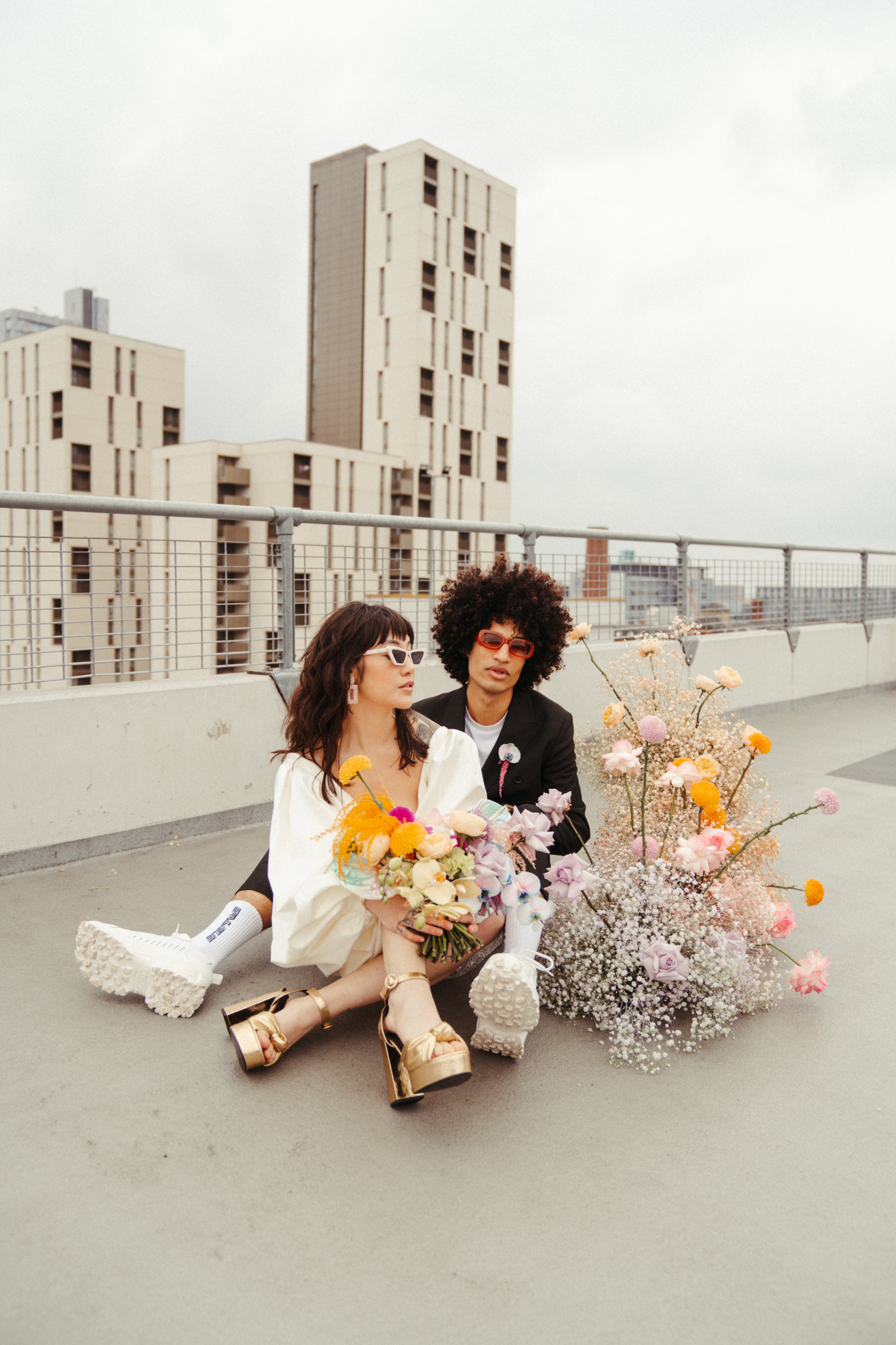  The bride and groom are sat on the floor next to each other on the top floor of an empty car park. Behind them are tall concrete buildings.   The bride is wearing a short white wedding dress with long voluminous sleeves, gold platform, open toed san