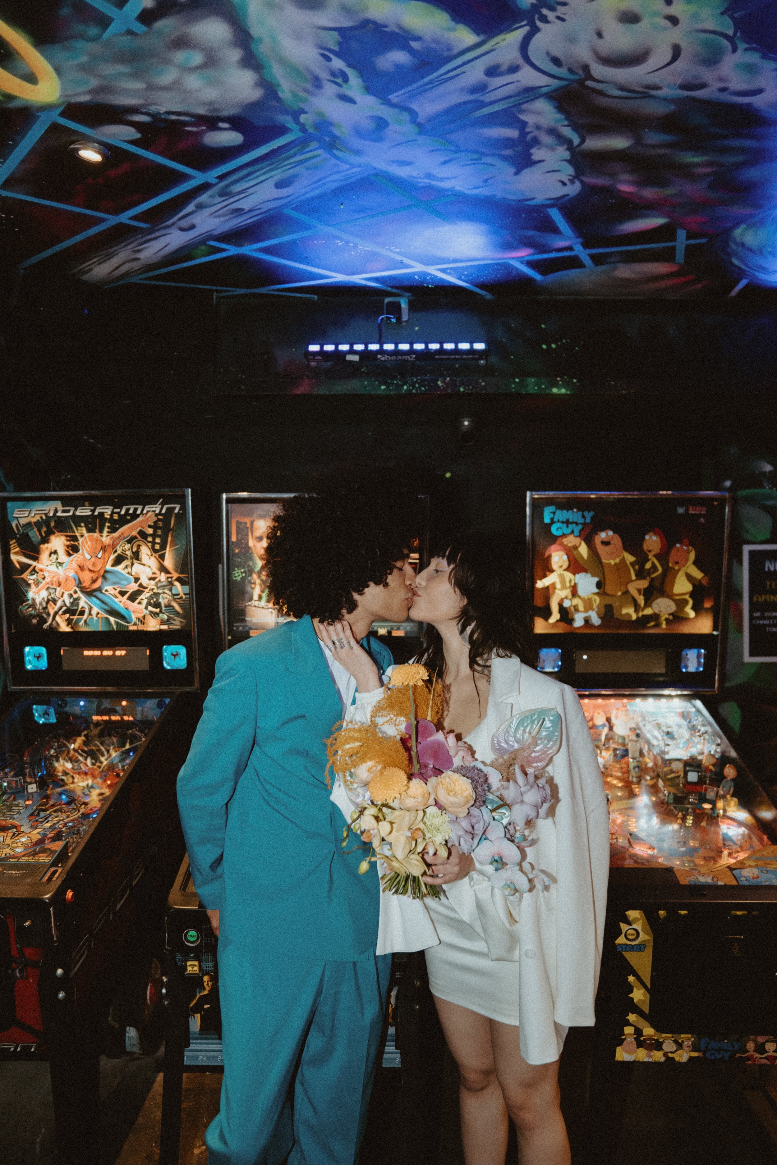  Bride and groom are stood in an arcade in front of pinball machines kissing. The bride is holding her hand tied wedding bouquet in front of her which is made up of various blooms with different textures in pale pinks, purples and creams with soft or