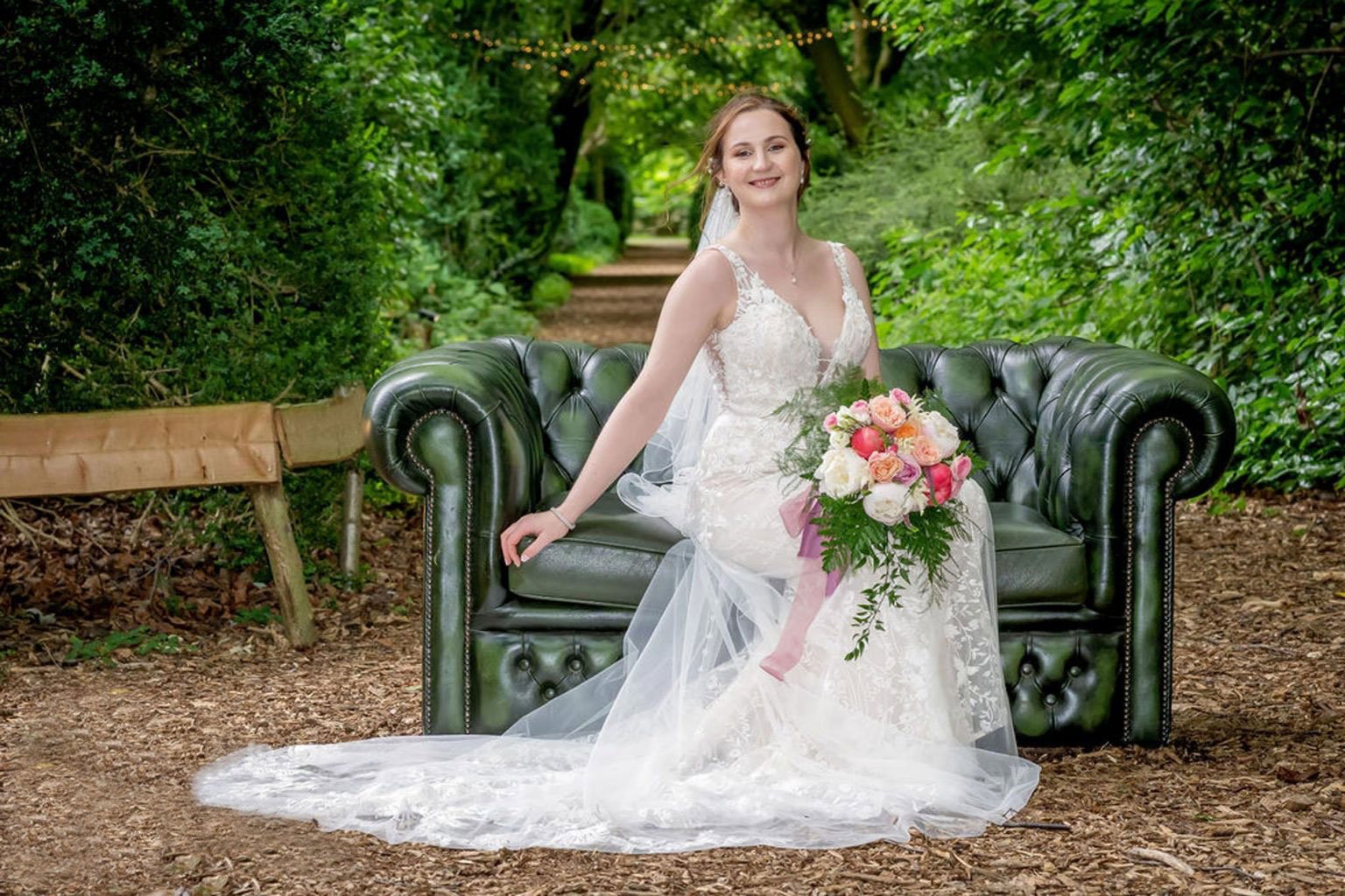  Bride sat on a green Chesterfield sofa which has been placed outside on a bark lined path with woodland either side.  The bride is holding her hand tied bridal bouquet in her lap. The various sized blooms in pinks and white are framed by green ferns