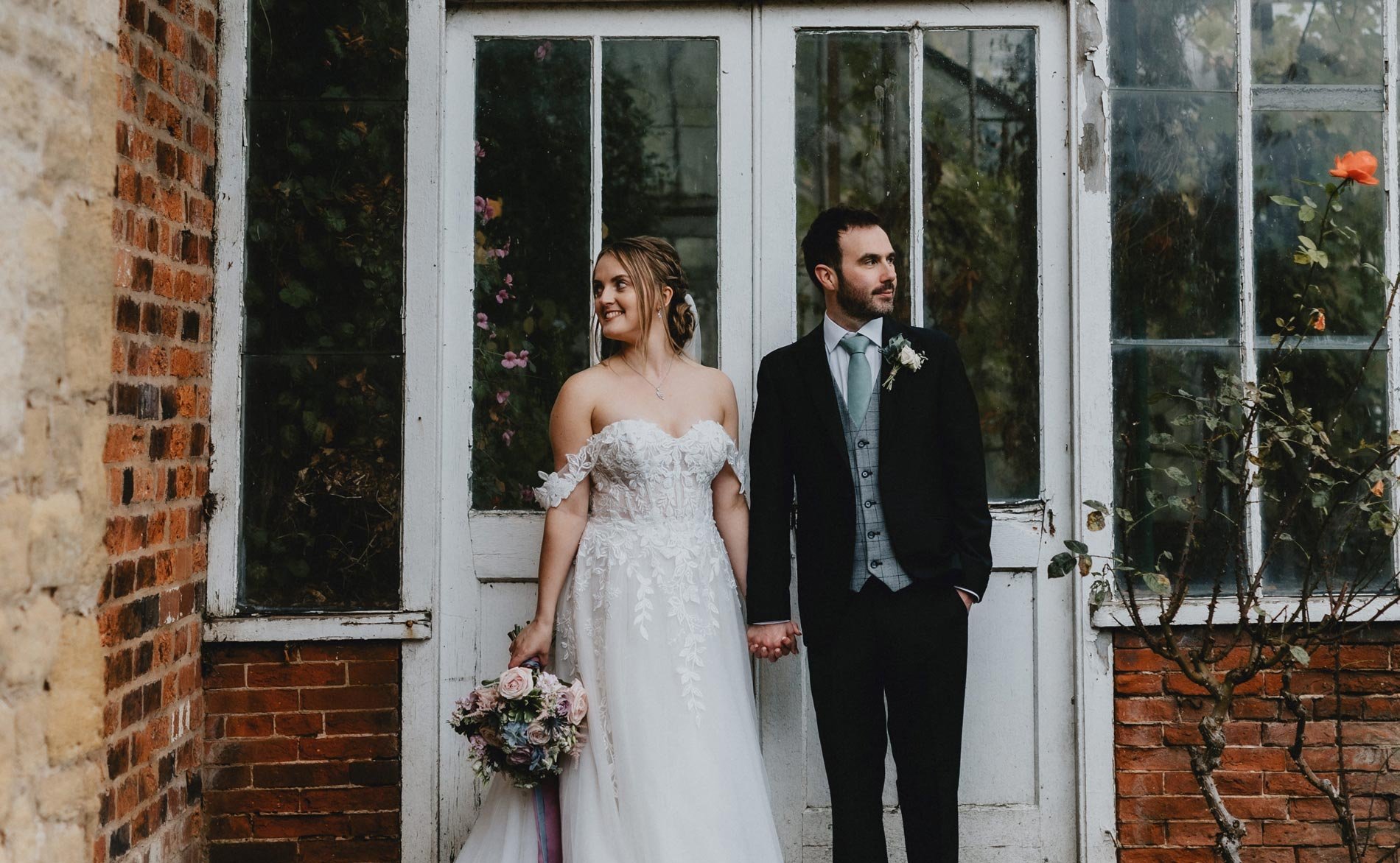  The bride and groom are stood outside holding hands looking in opposite directions to each other. Behind them are white weathered wooden doors into an orangery. The bride is holding her hand tied bouquet made up of pastel blue, pinks and white bloom