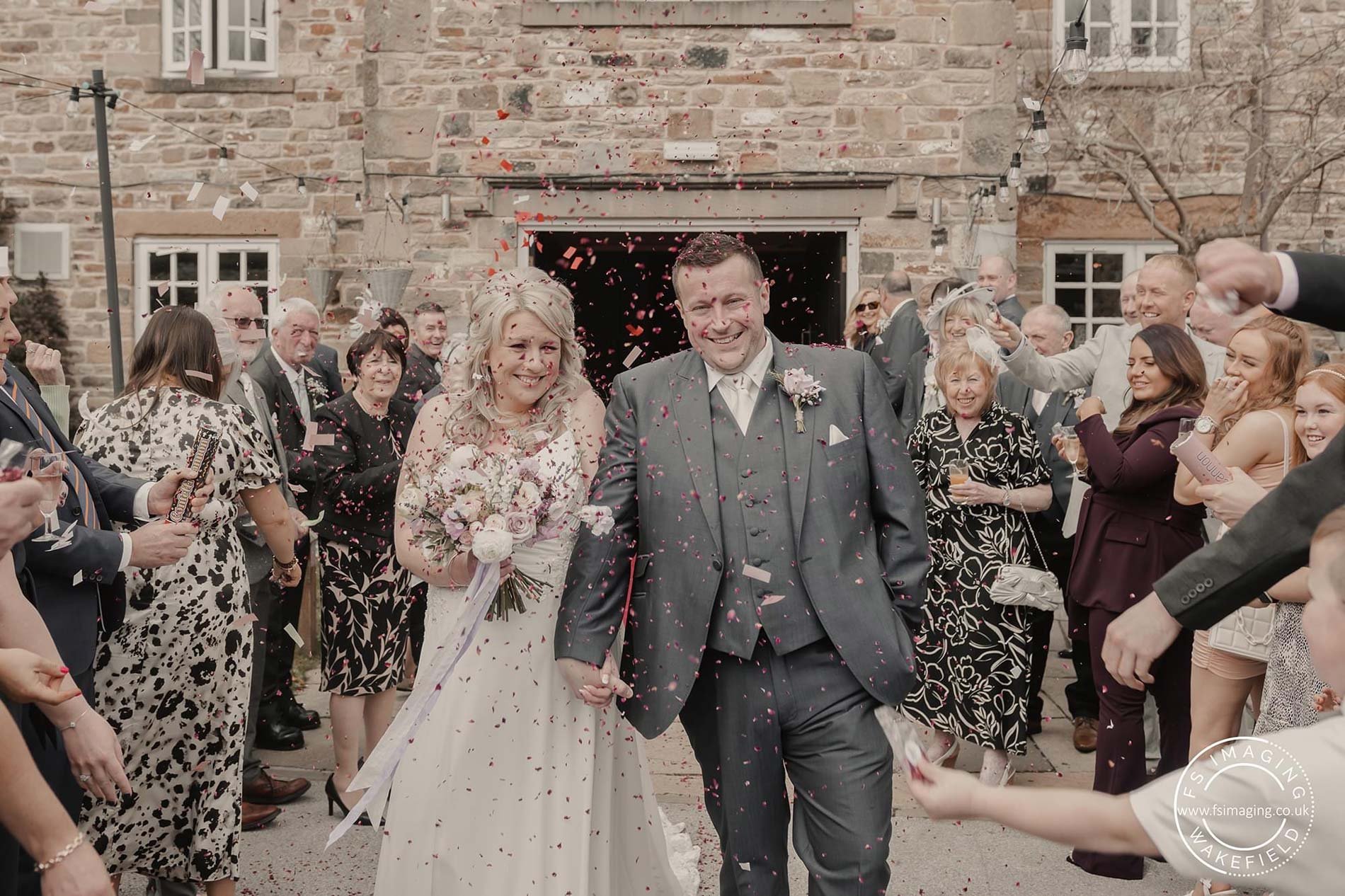  With a beautiful stone building behind them, the bride and groom are walking towards the camera with their wedding guests who are throwing confetti either side.   The bride is wearing a sleeveless, long white wedding dress and is holding her soft pa