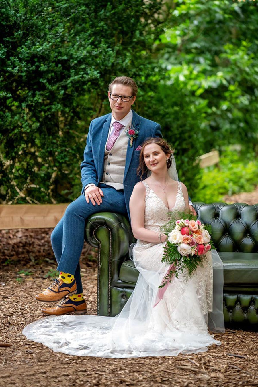  A bride is sat on a green Chesterfield sofa with the groom perched next to her on the arm. They are outside with green trees behind them.  The bride is wearing a v-neck, sleeveless white wedding dress and the train of the brides dress is trailing ov