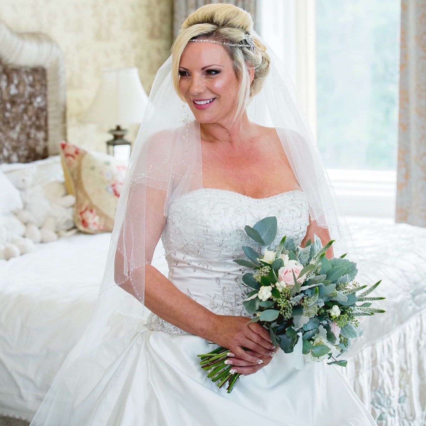  Bride wearing a long white, strapless wedding dress and white veil is sat on the corner of a bed made up with white bed linen. The bride is looking away from the camera holding her hand tied bouquet created with green foliage and white blooms.    Ph