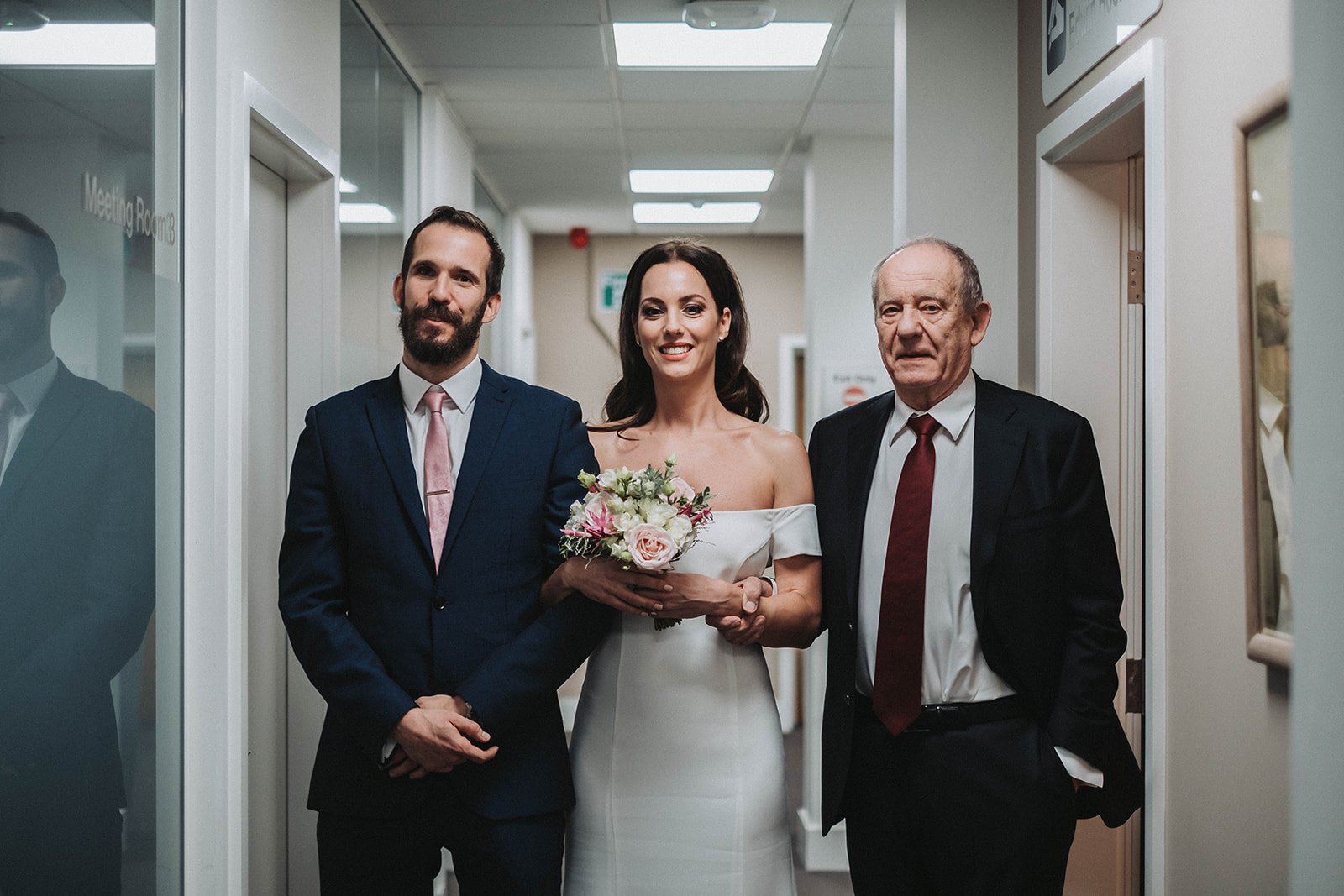  Bride is wearing a long, straight ivory, off the shoulder wedding dress, she is holding her small pink and cream hand tied bouquet in front of her. The bride is linked arms with a man on either side of her. The man on the left is wearing a blue suit
