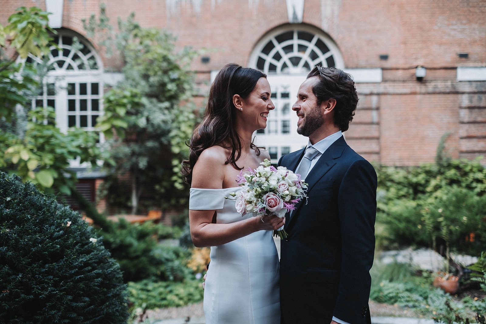  Bride and groom are stood outside amongst plants and shrubs with an impressive old building behind them. They are looking lovingly into each others eyes and smiling. The bride is wearing a sleek ivory wedding dress and the groom is wearing a blue su