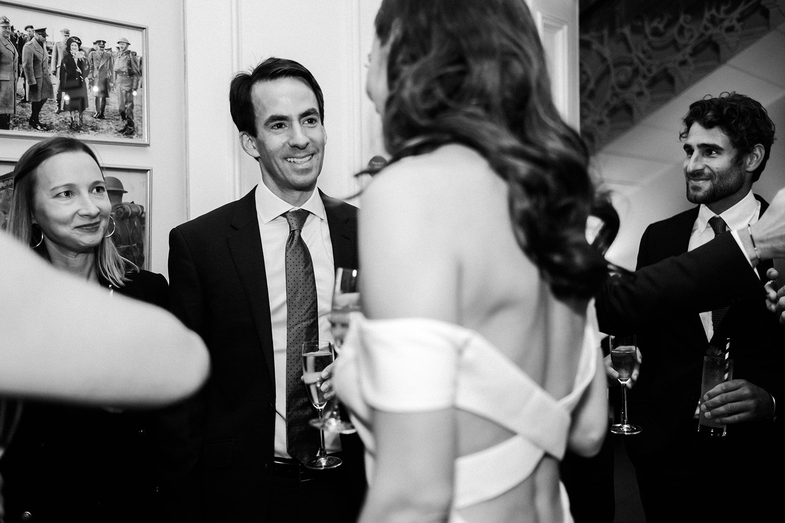  Wedding photographer has captured the bride engaging in conversation at the wedding reception with her wedding guests in this black and white image. You can see the back of the bride and her beautiful low cut, crossover back, sleek wedding dress.  