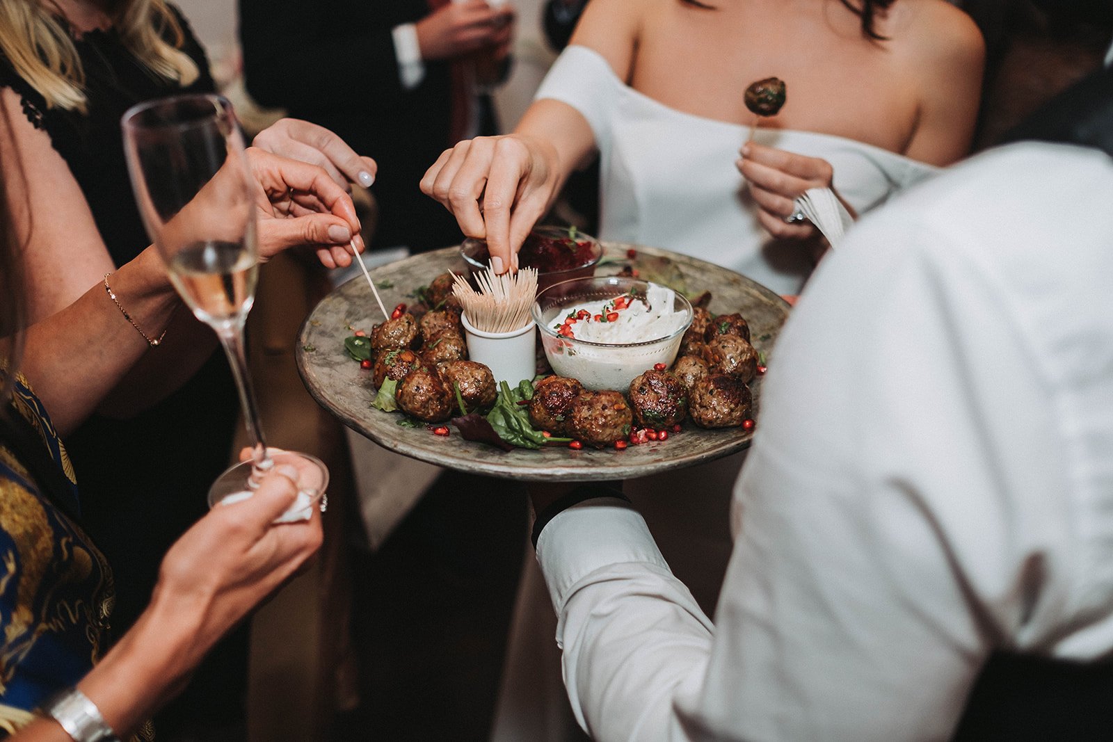  The wedding photographer has focused on the canapes which are being handed out by a member of the waiting staff wearing a crisp white shirt, The bride is reaching for a cocktail stick from the pot which has been placed amongst the meatballs and othe