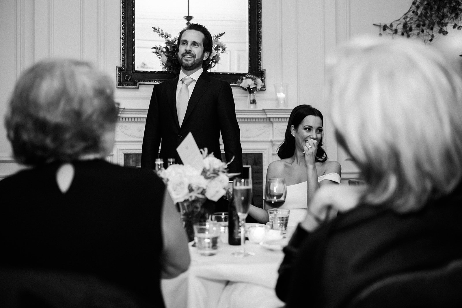  Black and white image of groom stood up giving a speech. The photographer has taken the photo through a gap between the wedding guests who are blurred in the foreground of the image looking towards the bride and groom. The groom is stood next to the