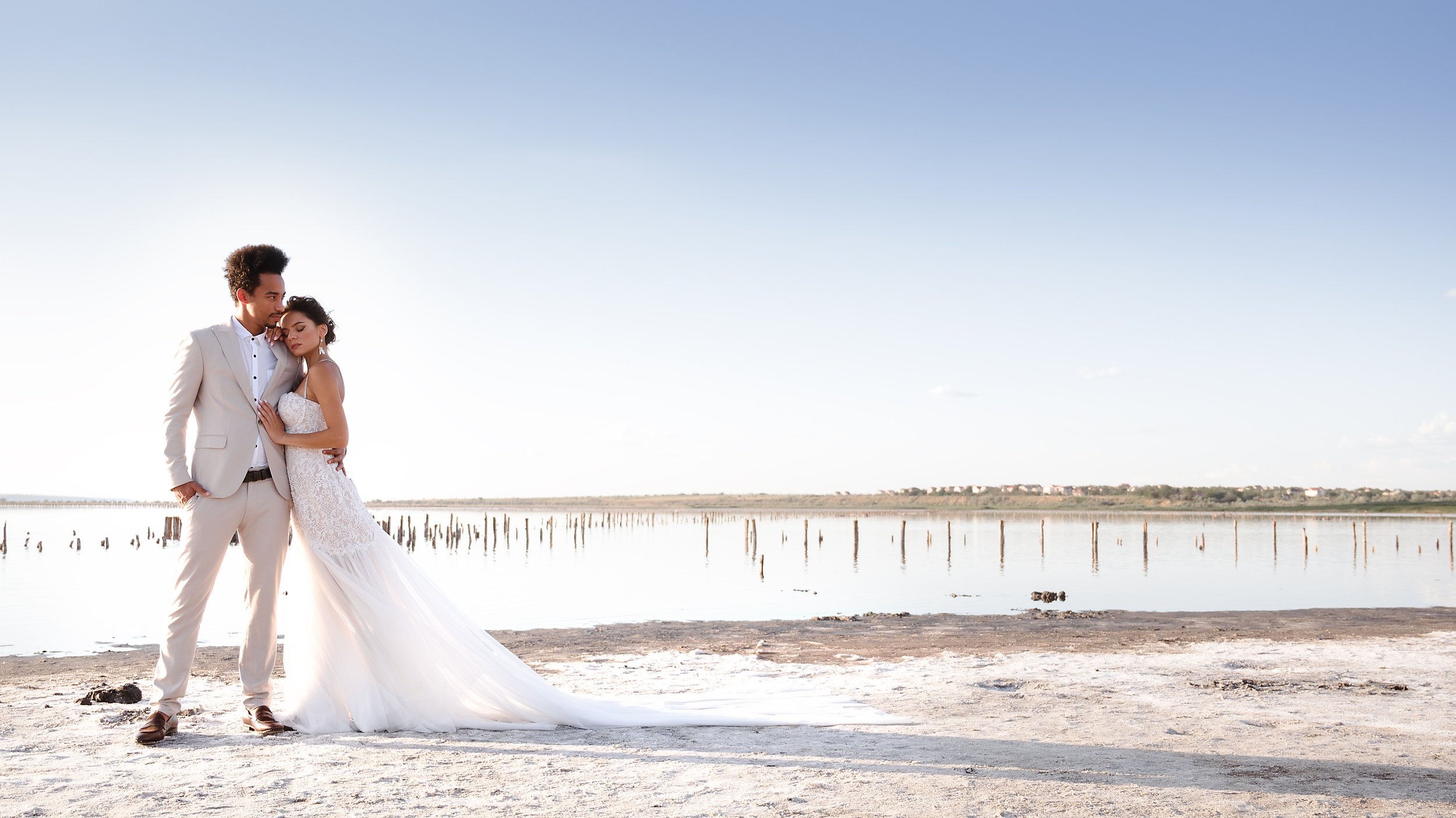  Under a clear blue sky the bride and groom are stood on the beach. The bride is leaning into the groom with her head resting on his shoulder and the groom has his left arm around the bride and is other hand in his pocket.  The groom is wearing a cre