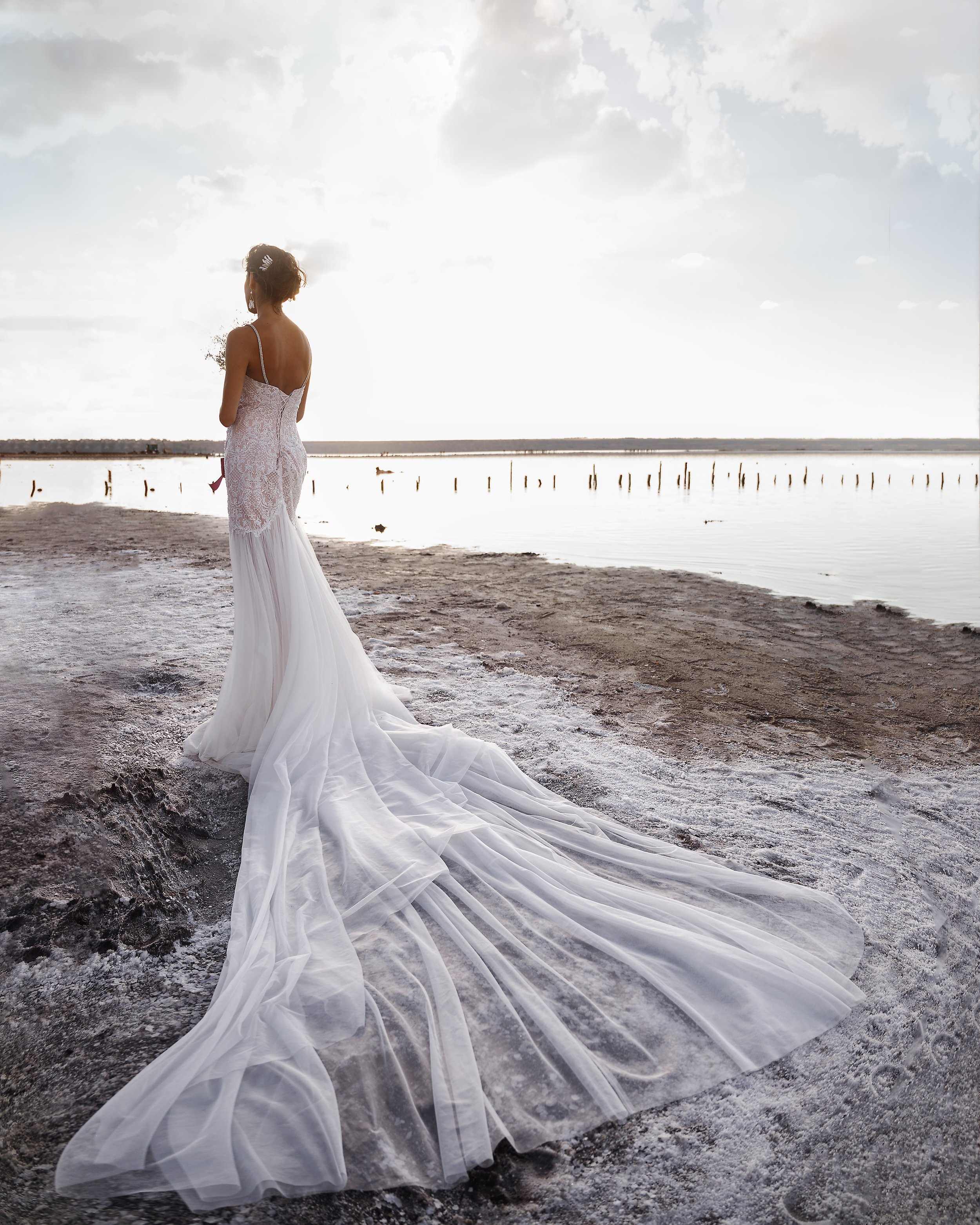  Under a grey cloudy sky, the bride is stood on a beach with her back to the wedding photographer looking out to the distance across the sea. The bride is wearing a white, strappy, long wedding dress with a long train which has been laid out across t