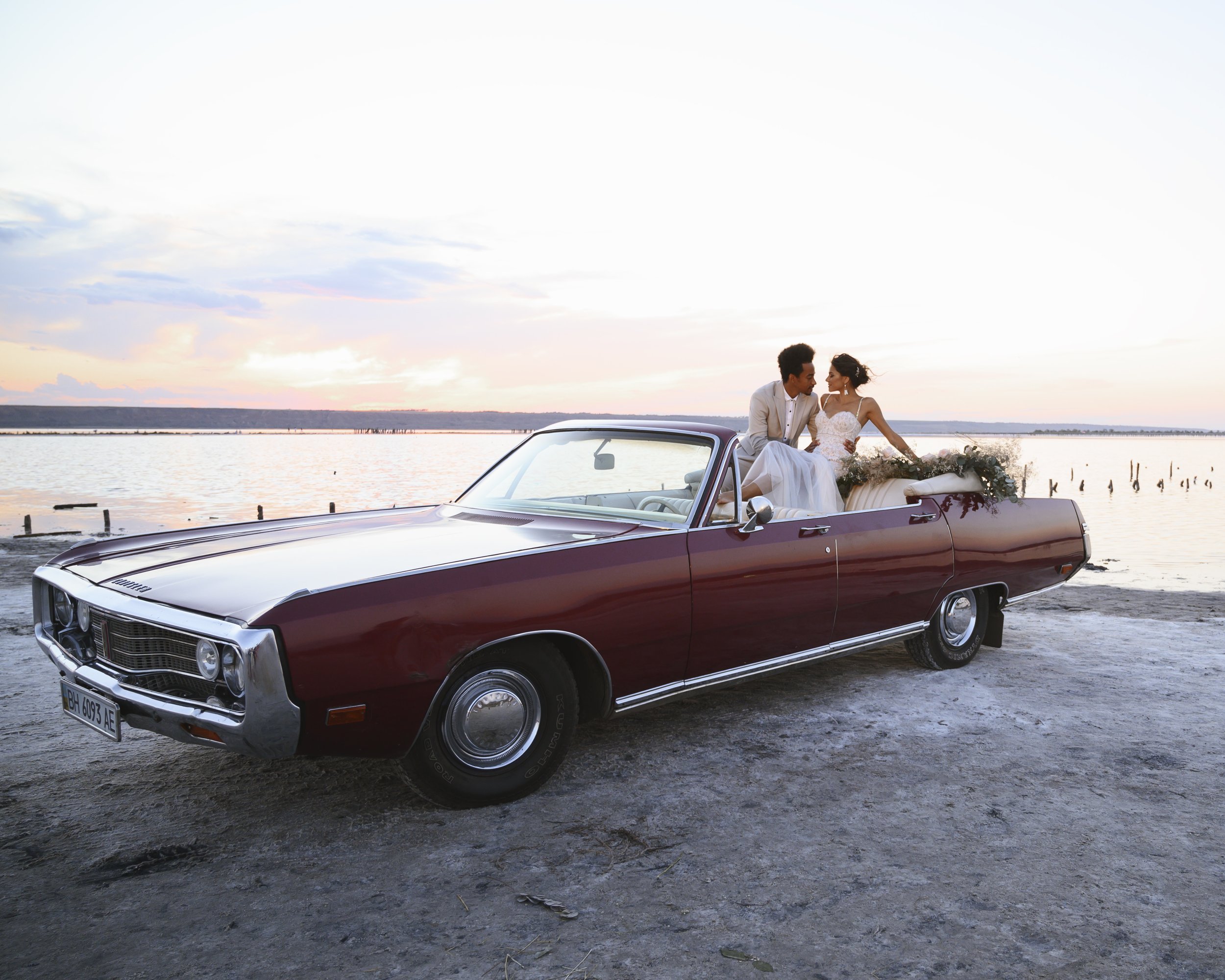  A red convertible car with cream leather interior is parked on a beach. With the sea and sunset behind them the bride and groom are sat on the top of the back seats with their feet resting on the backs of the front seats and are looking into each ot