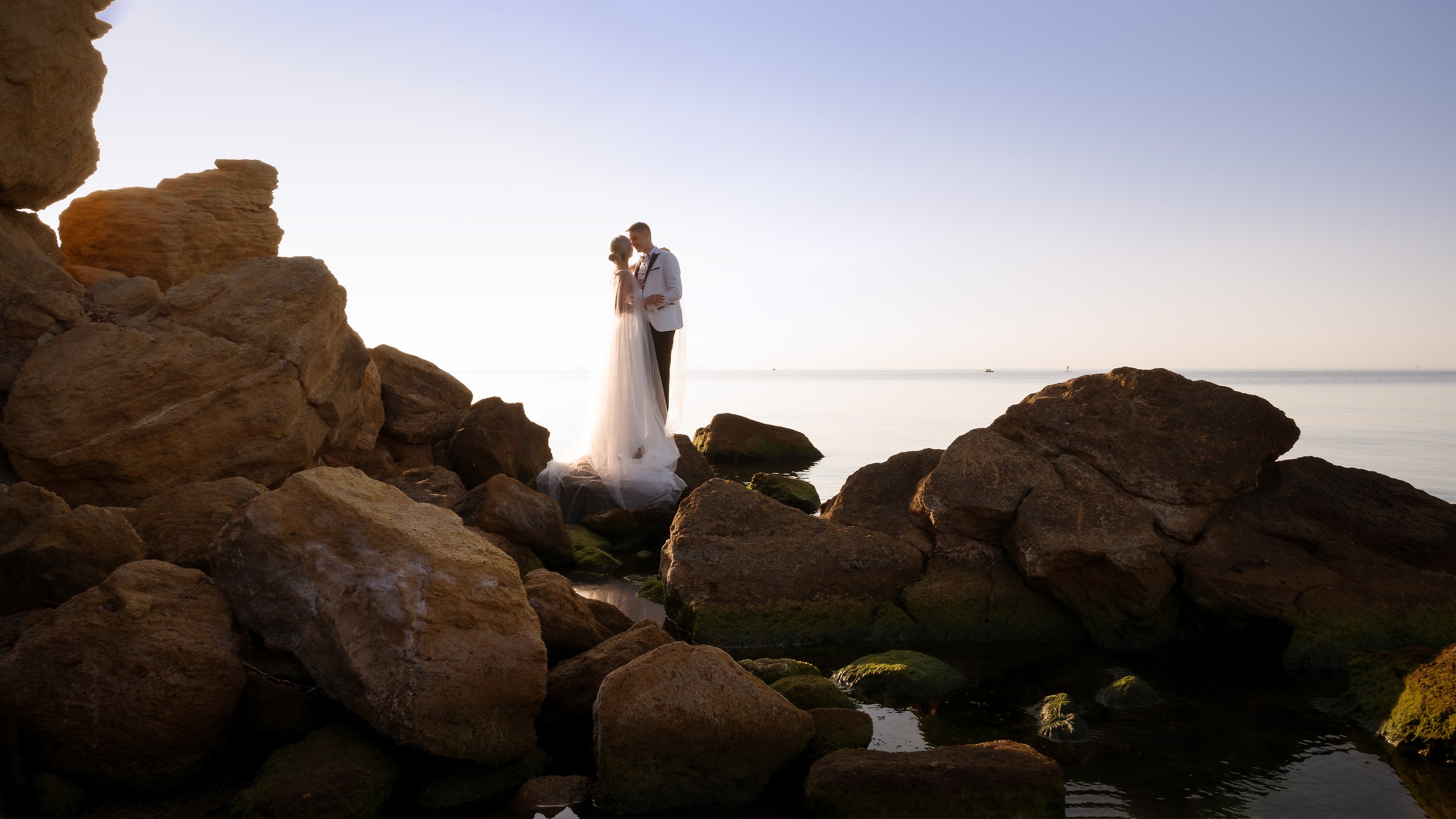  With nothing on the horizon and under a clear blue sky, the wedding photographer has captured the bride and groom stood out on rocks in the sea. The couple are facing each other looking into each others eyes, The groom has his arms around the brides