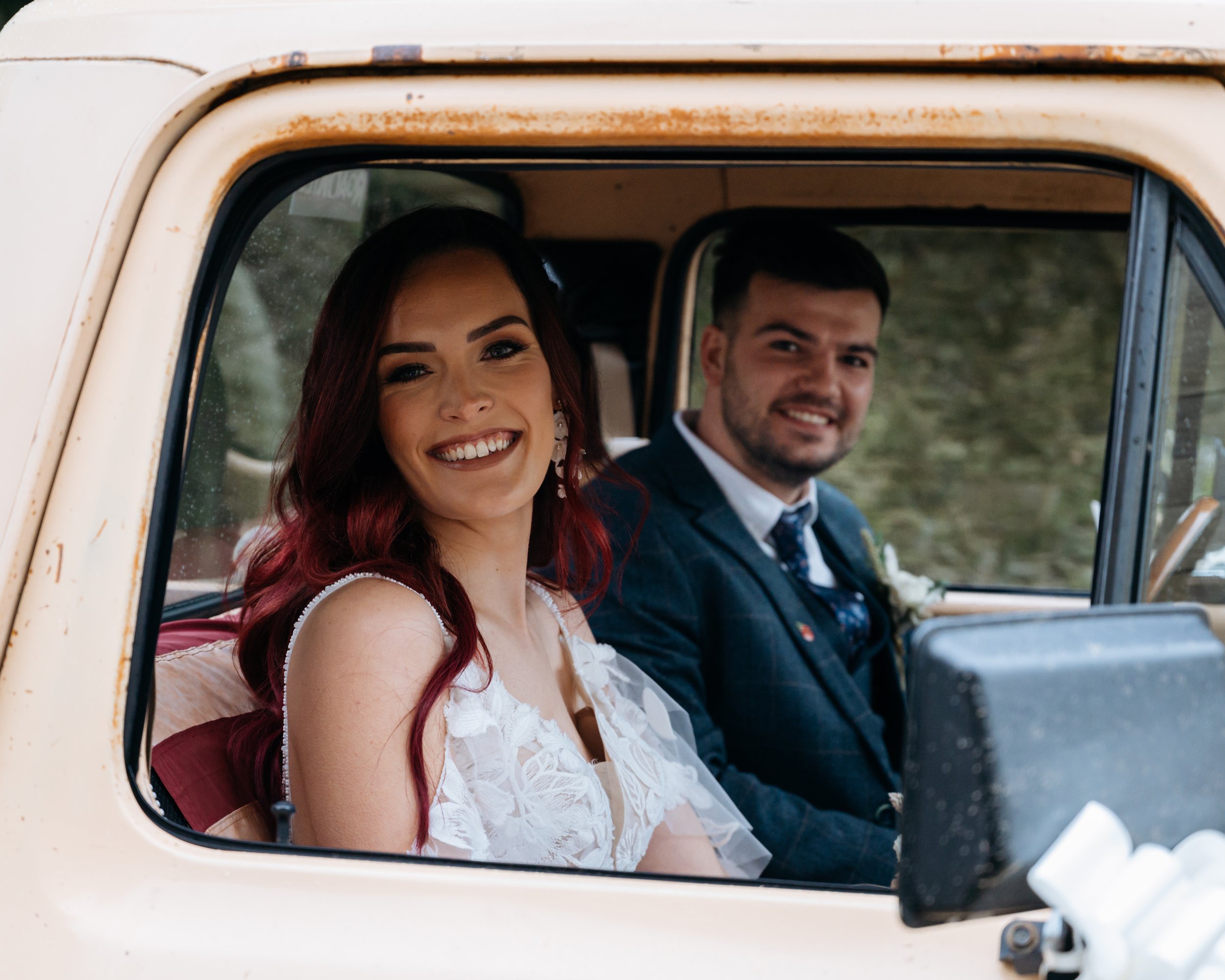  With the groom at the wheel, the wedding photographer has taken this photo of the bride and groom sat in the cab of a beige pickup truck. The groom is wearing a navy 3-piece suit with and a brown check stripe and the bride is wearing a strappy white