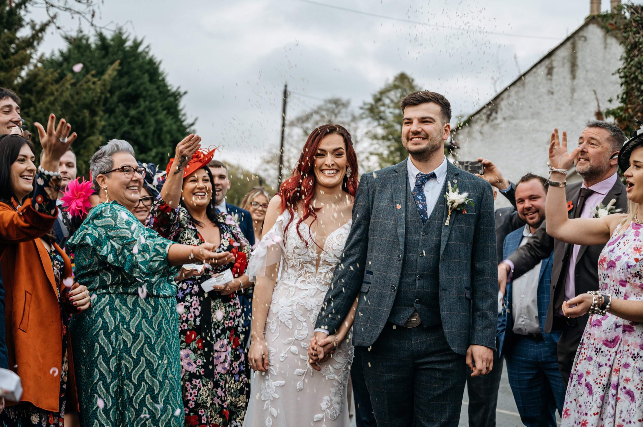  The bride and groom are walking outside towards the wedding photographer holding hands and smiling. The wedding guests on either side are throwing confetti over them. The groom is wearing a navy and brown check 3-piece suit with a navy floral tie an