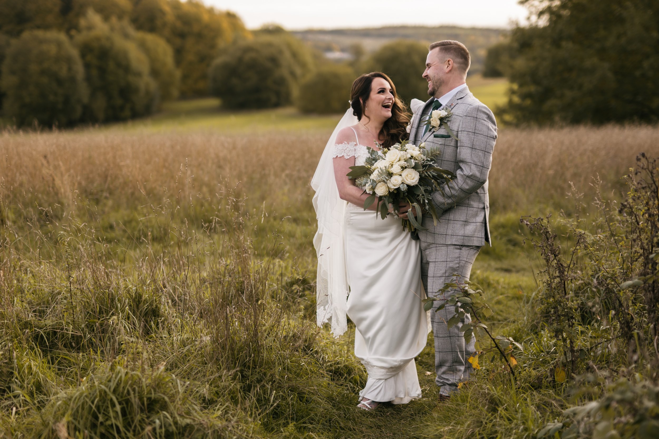  In a field of long grass the wedding photographer has captured this natural image of the bride and groom sharing a special moment of laughter. The couple are walking closely next to each other both with a hand holding the bridal bouquet, whilst look