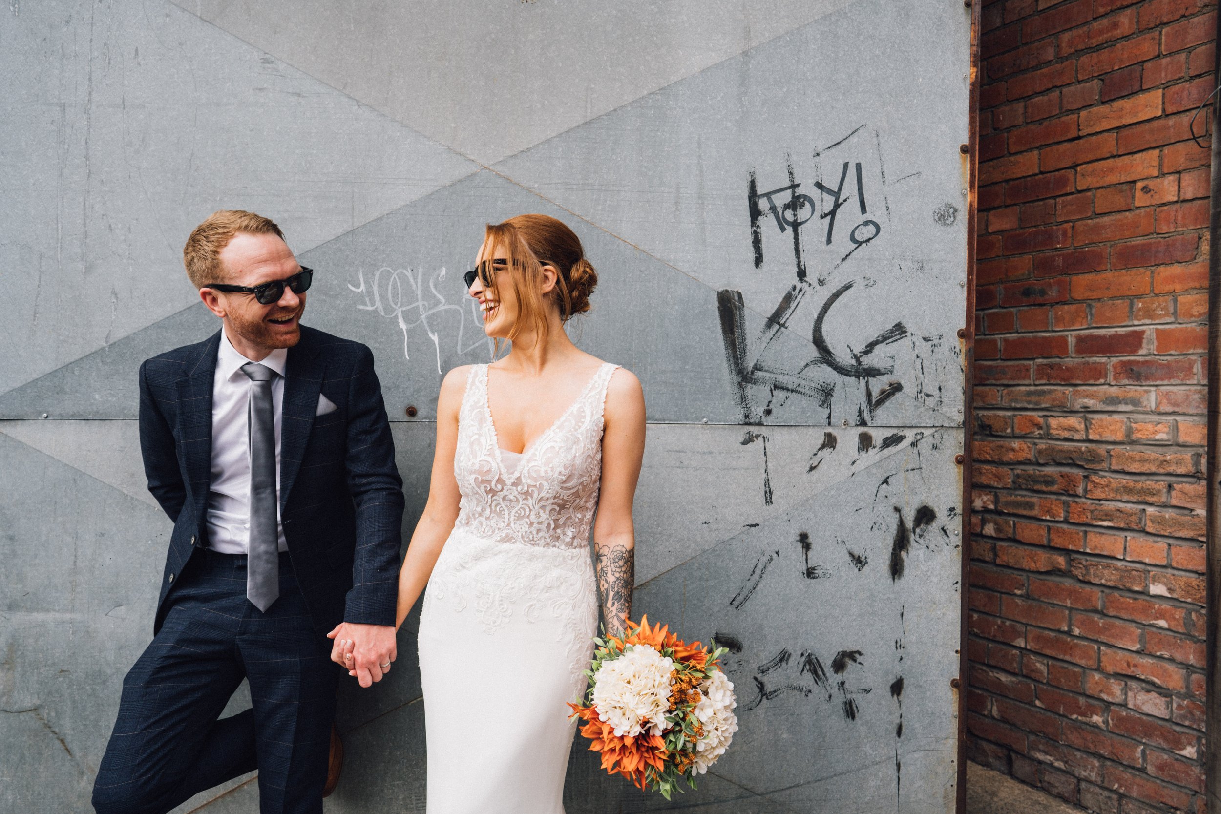  Photographer Credit: Alexandra Holt Photography  Bride and groom are stood outside in front of a grey concrete wall with black graffiti on it. They are holding hands looking at each other and laughing whilst both are wearing sunglasses. The groom is