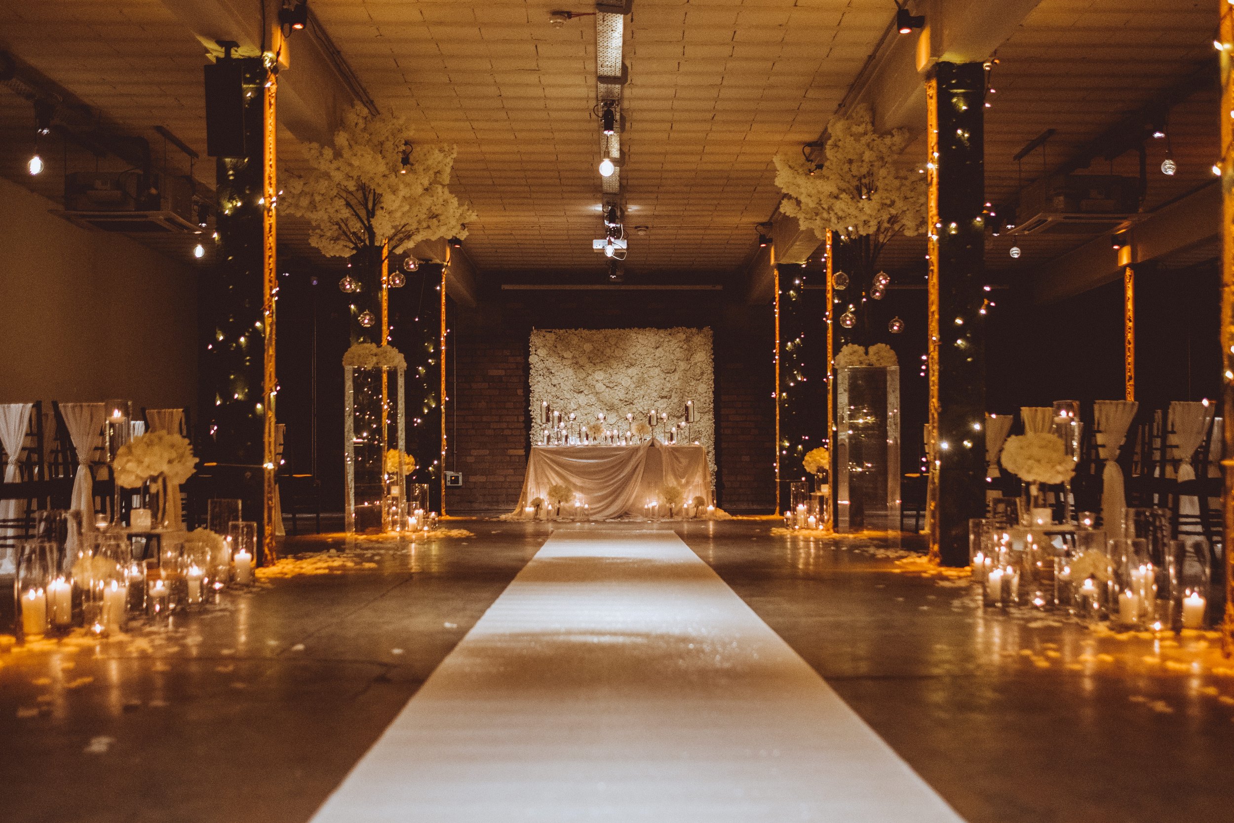  Photographer Credit: Dorian Knightley  A beautiful view of the wedding aisle leading up to the alter, set up inside the Victoria Warehouse.   There is a long cream runner on the floor down the aisle with pillar candles in storm vases grouped togethe