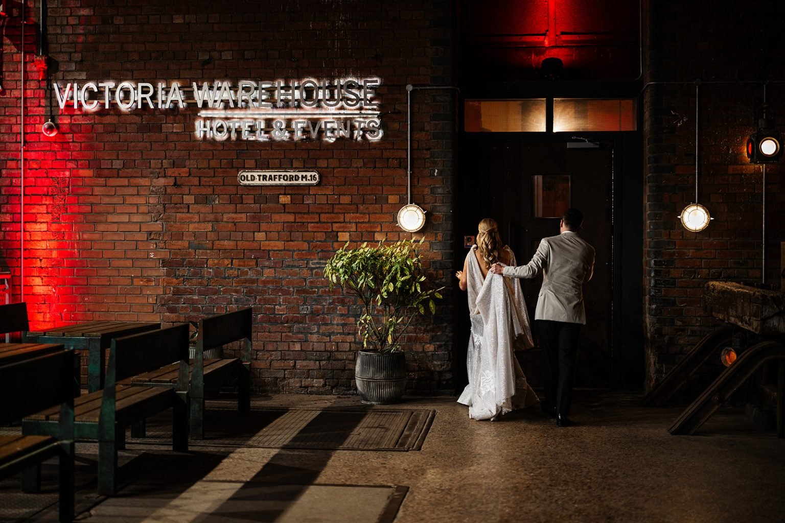  Photographer Credit: Hollie &amp; Patrick Mateer  Bride and groom about to walk through a doorway inside the beautiful Victoria Warehouse. To the left of the bride there is an exposed brick wall with VICTORIA WAREHOUSE sign lit up and underneath it 