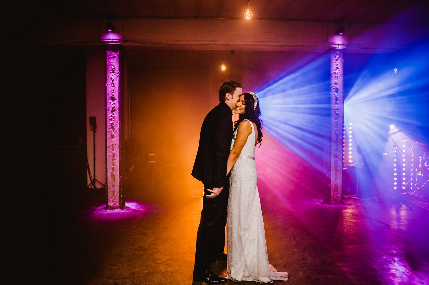  Photographer Credit: Kate McCarthy  In a beautifully lit room within the Victoria Warehouse the bride and groom are stood close together with the groom’s face behind the bride’s as he is whispering in her ear. The large room is empty except for the 