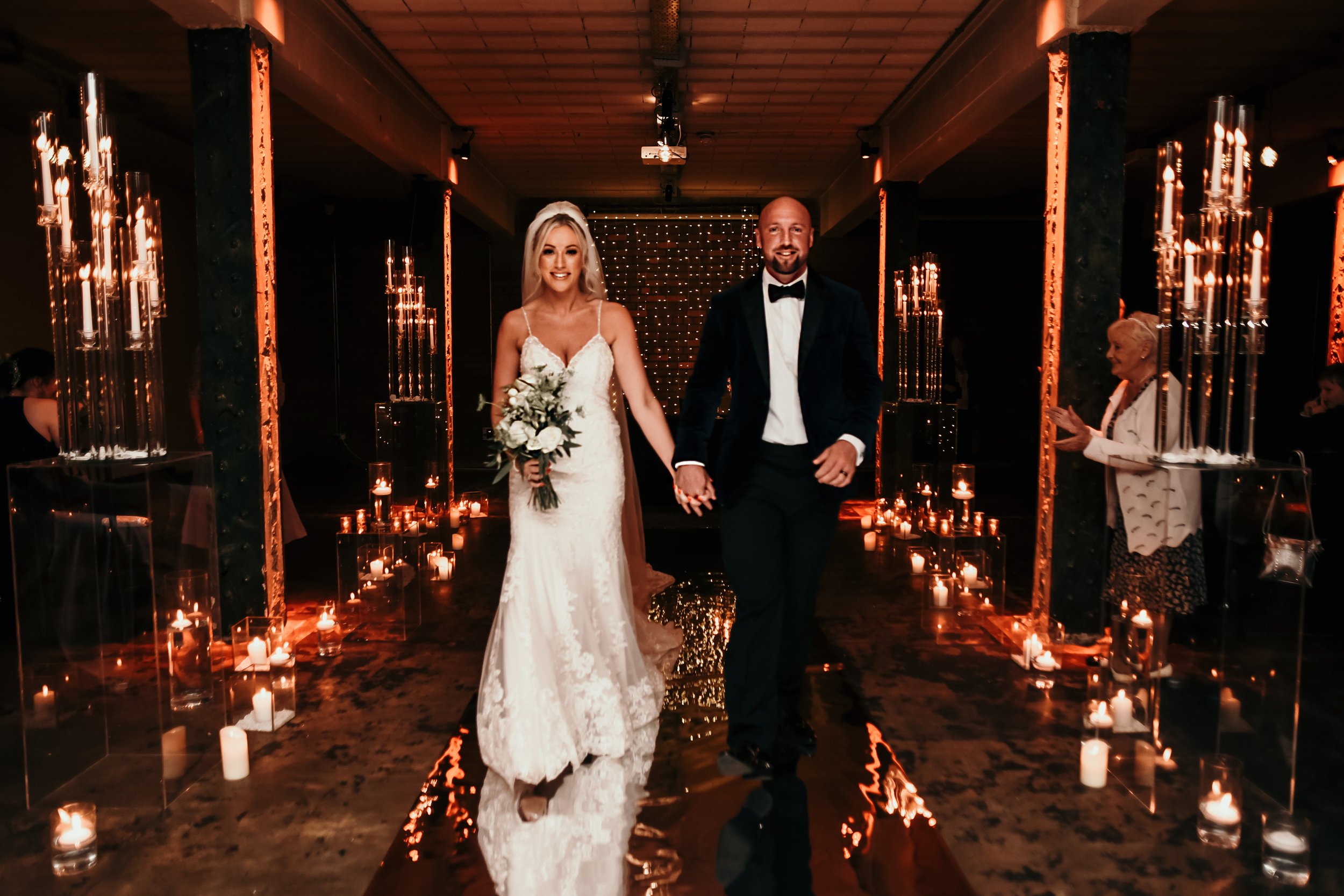  Photographer Credit: Kerry Lu  Bride and groom have been photographed walking back up the aisle within the stunning Victoria Warehouse venue. The room has been dressed with a copper panel down the aisle for the couple to walk on and either side ther