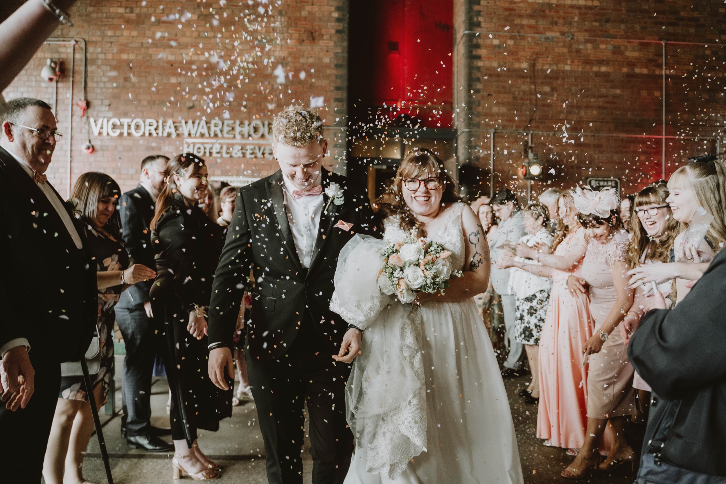  Photographer Credit: Kev Jacutan  Inside a bright exposed brick room within Victoria Warehouse the bride and groom are walking back up the aisle with their wedding guests either side of them throwing pastel confetti.  The bride is wearing a long, cr