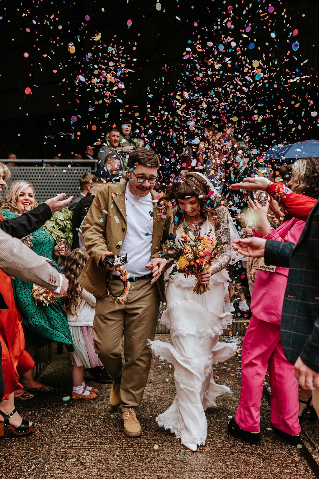  Photographer Credit: Maddy Farris  Outside the beautiful Victoria Warehouse in the heart of Manchester city centre the bride and groom are walking through a shower of bright confetti which is being thrown by their wedding guests who are lining the p