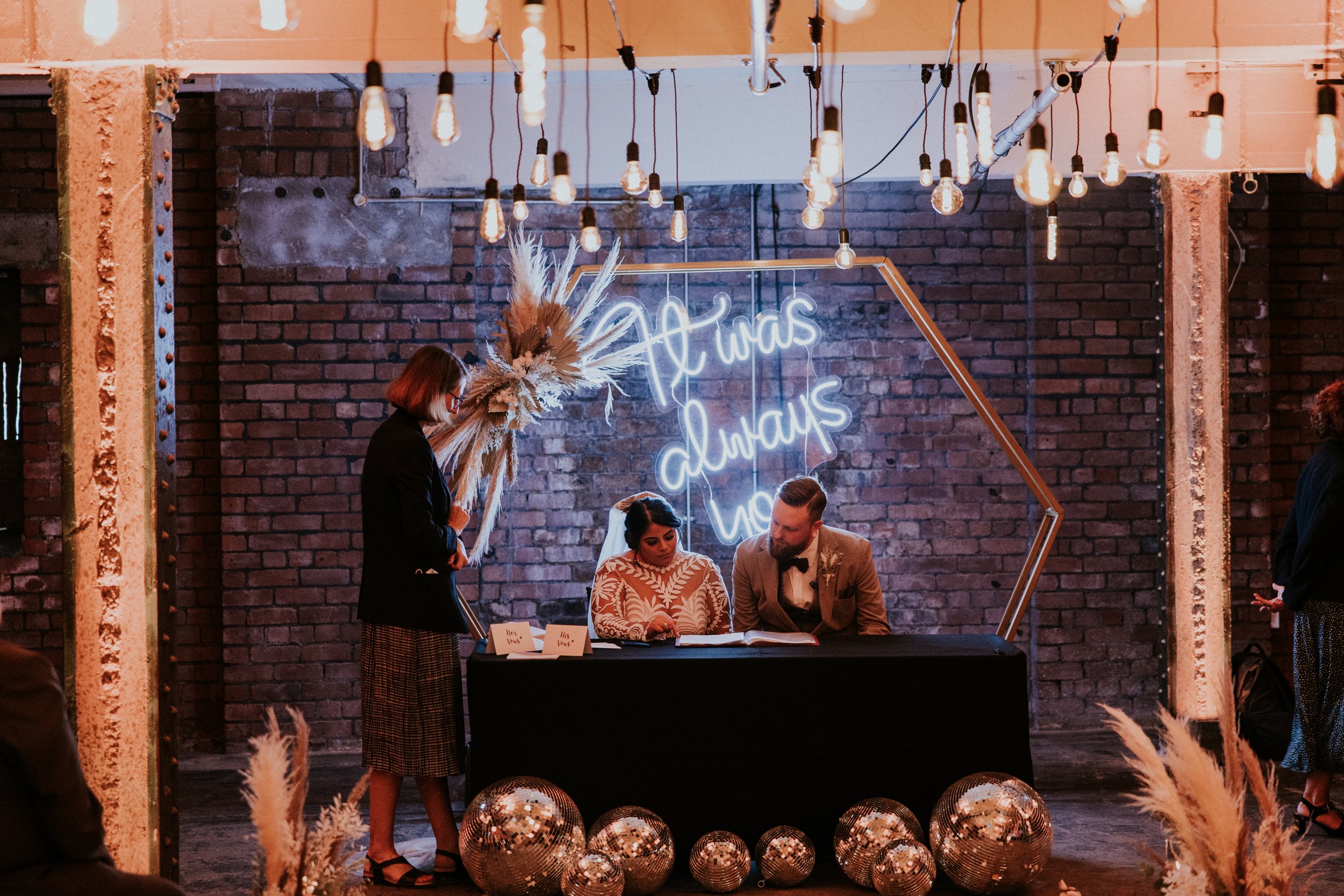  Photographer Credit: James Morris Photography  Inside Victoria Warehouse wedding venue. Bride and groom are sat at a table signing the wedding register. The table is covered in a black cloth with various size mirror balls along the floor. There are 