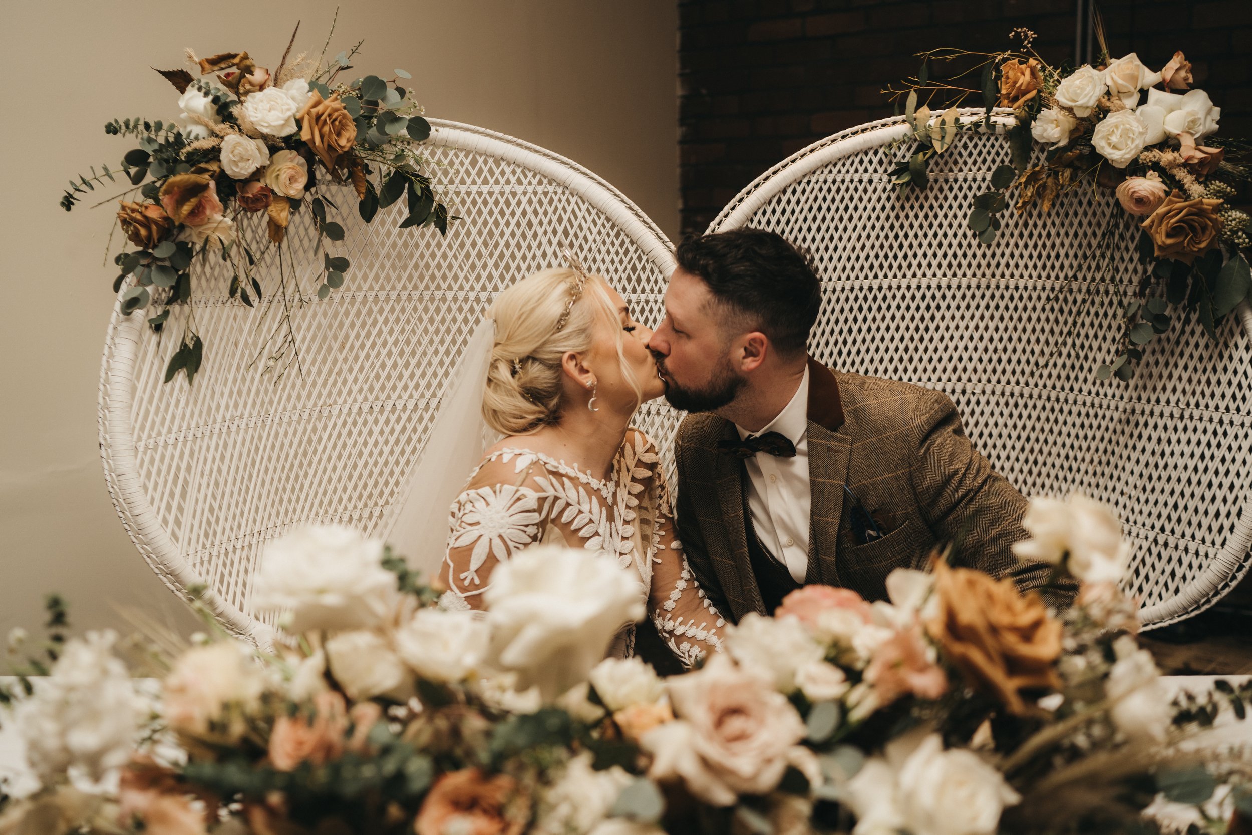 Photographer Credit: Mariola Zolads Photography  In Victoria Warehouse, the bride and groom are sat on large white wicker chairs sharing a kiss.   A floral installation on the table in front of the couple is made up of creams, pinks and greens and i