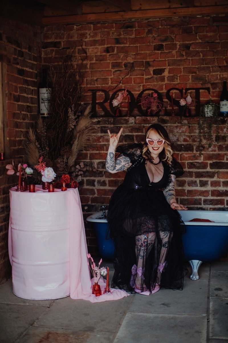  Photo Credit: Alt Wedding Co   The wedding singer &amp; guitarist is wearing a long lacy black dress with short sleeves and sunglasses. She is sitting on the side of a freestanding blue bath whilst holding her right hand up  with her fingers forming