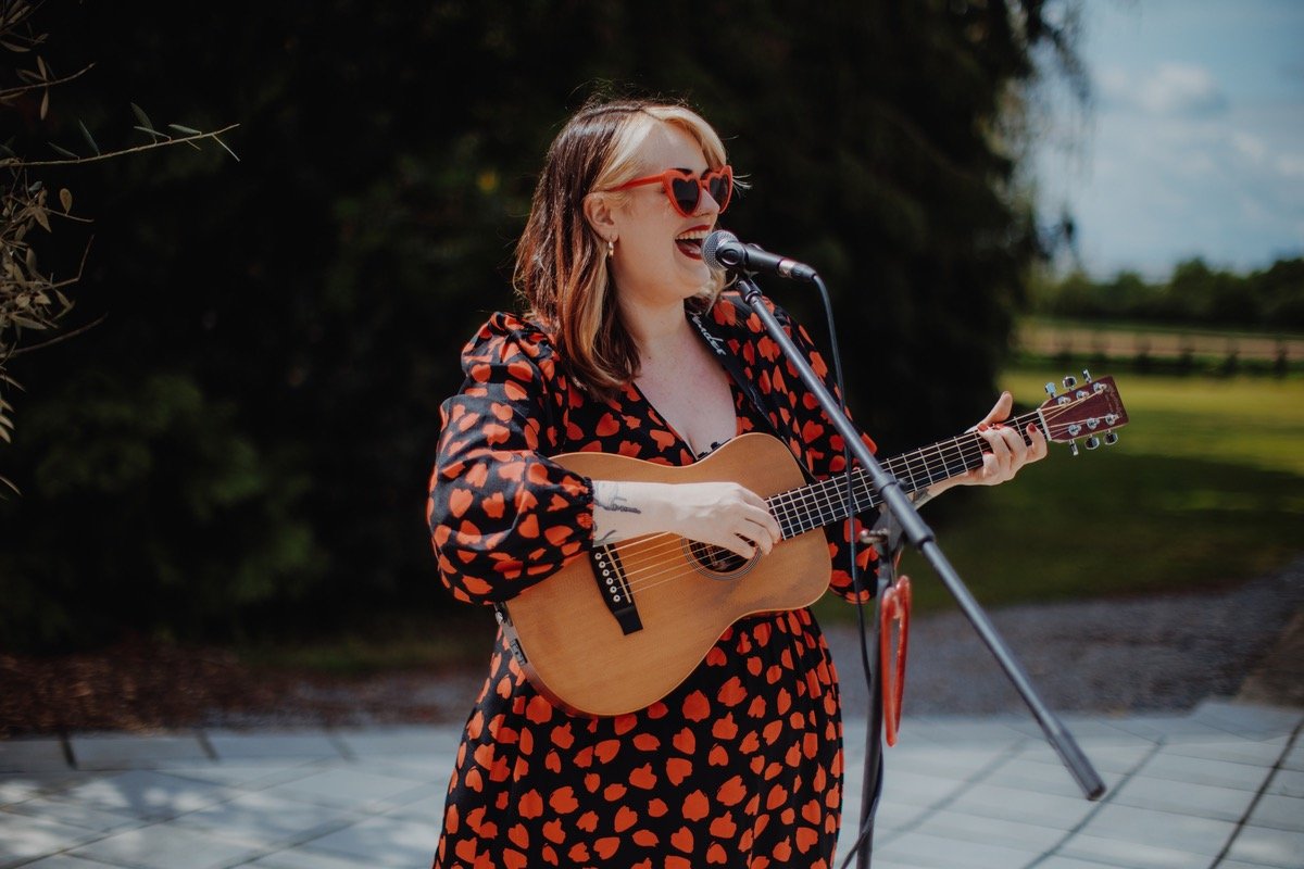  Photo Credit: Alt Wedding Co   Playing her guitar and wearing a long black dress patterned with red hearts and red heart sunglasses, the wedding singer &amp; guitarist is performing outside.  