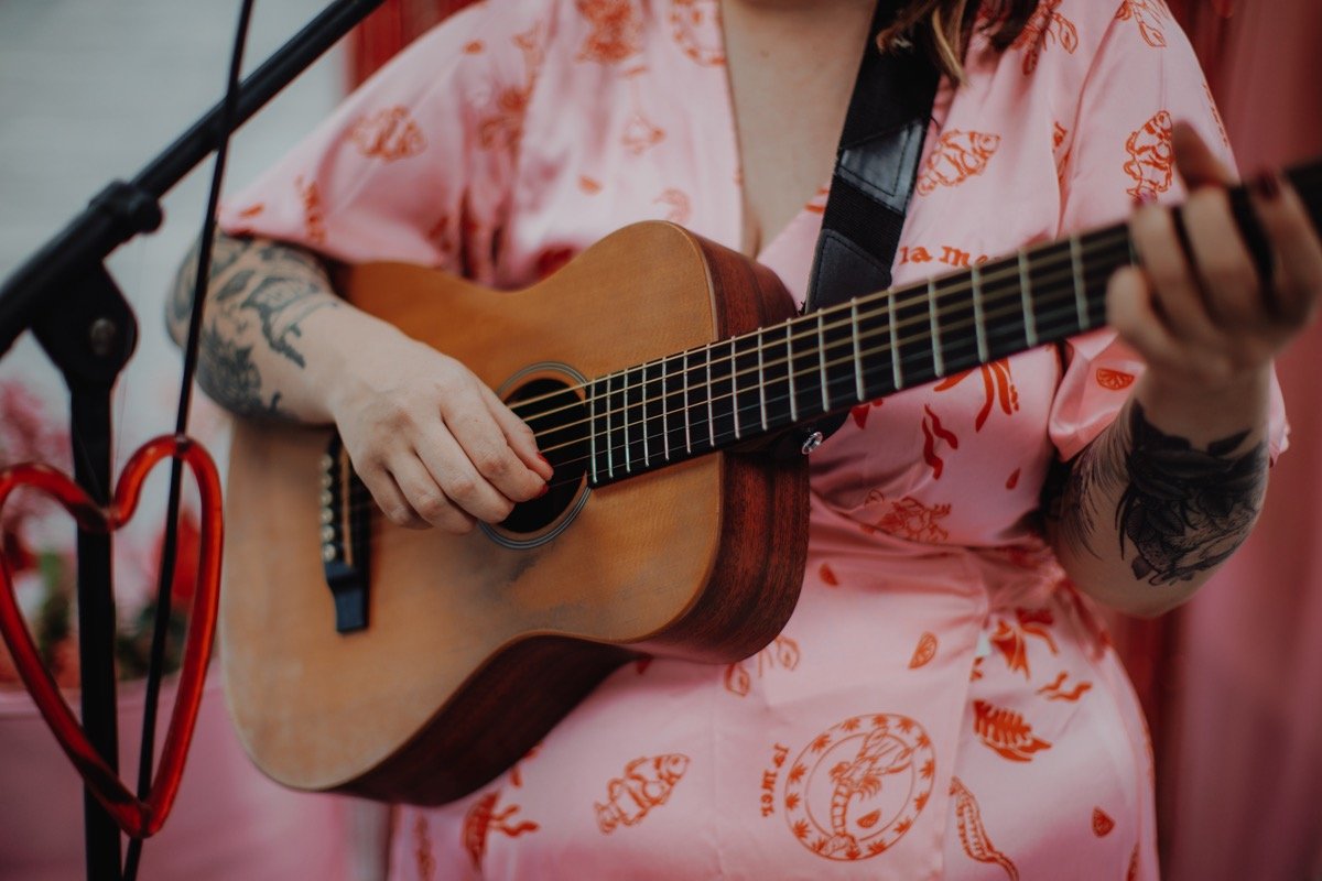  Photo Credit: Alt Wedding Co   An upclose image of the wedding entertainer playing her acoustic guitar. A red heart hangs from the microphone stand. The wedding singer is wearing a short sleeved, long pink wrap dress with red fish and sea print patt