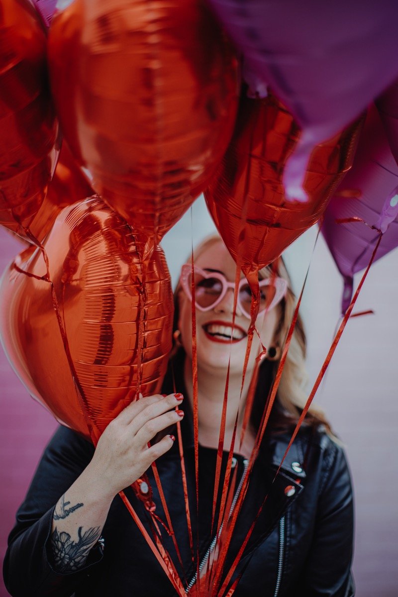  Photo Credit: Alt Wedding Co    The wedding singer can be seen through the red strings of the red and purple heart balloons she is holding. She is wearing a black biker jacket and pink heart sunglasses and is looking up at the balloons in front of h