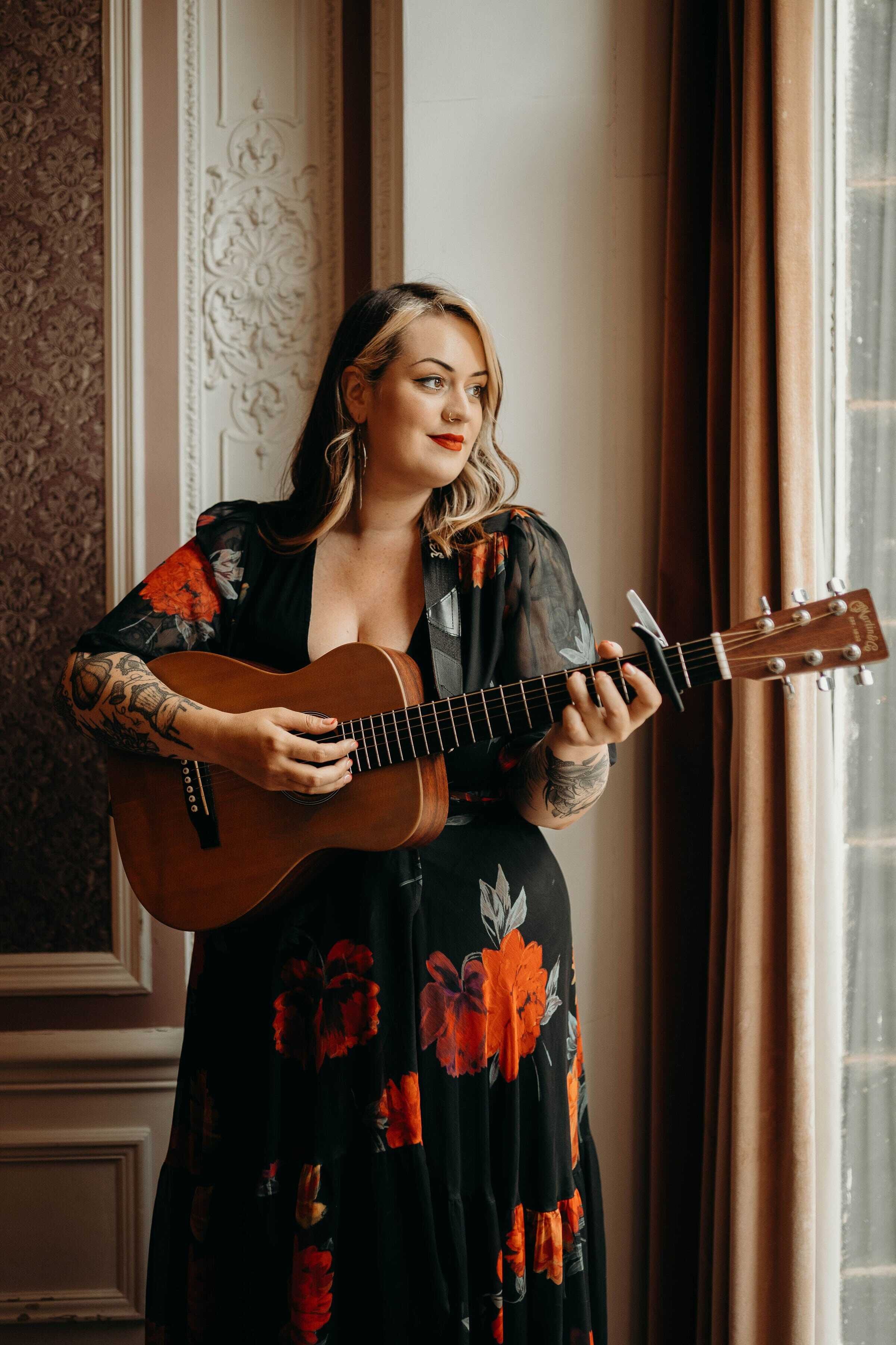  Photo Credit: Charlotte Jane Photography  Wedding singer and guitarist is stood in a grand room looking out of a window whilst playing an acoustic guitar. She is wearing a long dress with short sleeves  which is black with large red flowers printed 