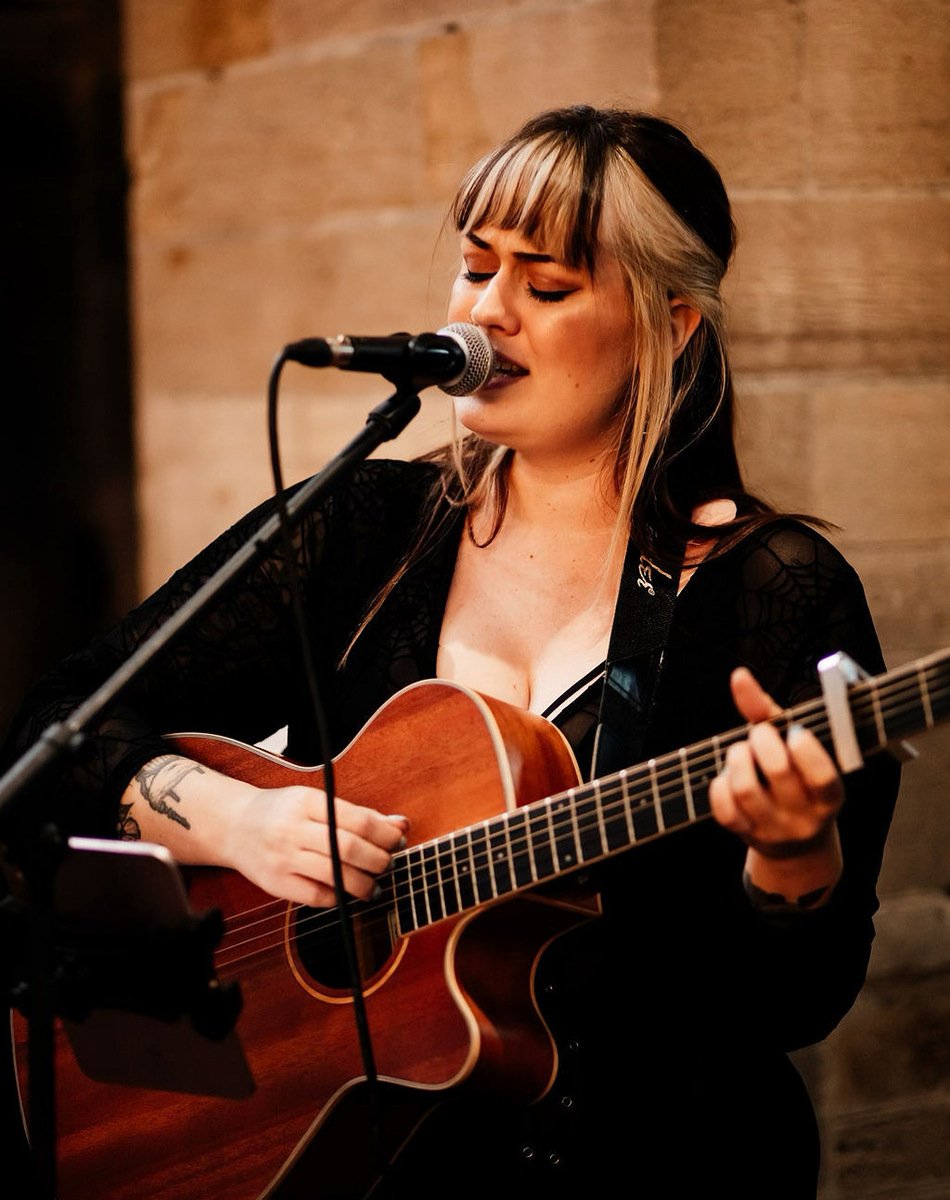  Photo Credit: Gem Wright Photography  The performer has been captured in the moment with her eyes closed during a wedding ceremony. She is singing into the microphone which is on a stand and playing an acoustic guitar. She is wearing a long sleeved 