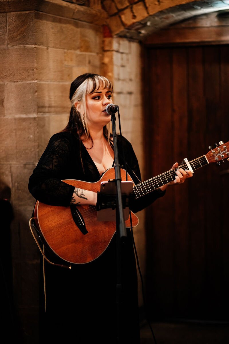  Photo Credit: Gem Wright Photography  Stood inside an old building with exposed stonework behind her and a dark wooden door to the side, the wedding singer and guitarist is captured performing looking out to the wedding guests. She is wearing a long